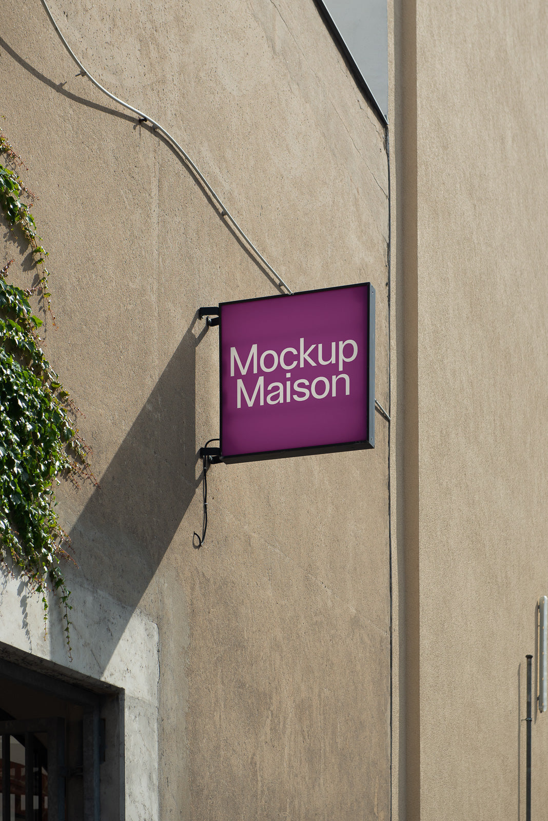 PSD mockup of a wall-mounted square sign with "Mockup Maison" text, displayed on a textured building exterior with green ivy.