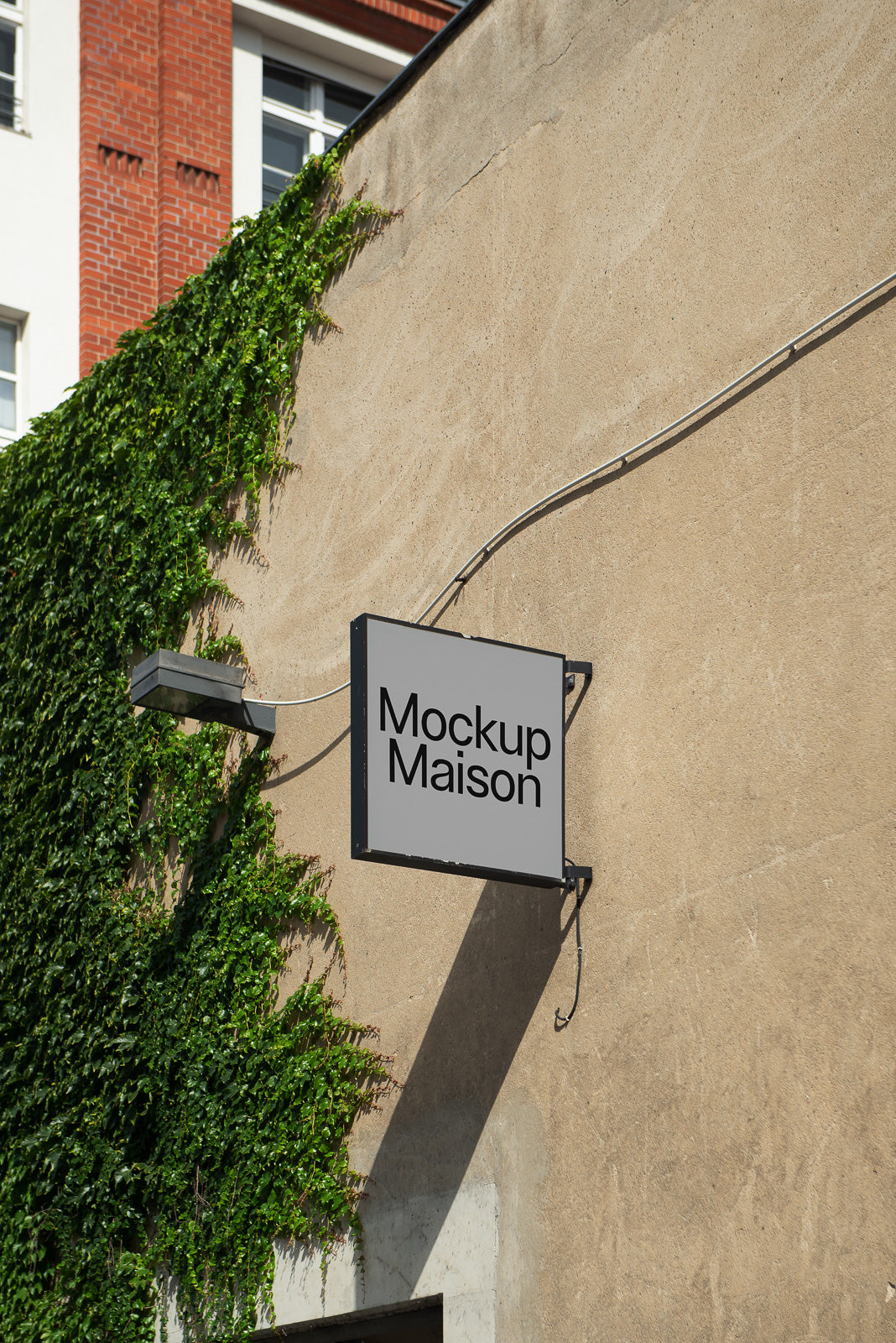 PSD mockup of a rectangular wall-mounted sign with "Mockup Maison" text, placed on a concrete wall partially covered by green ivy.