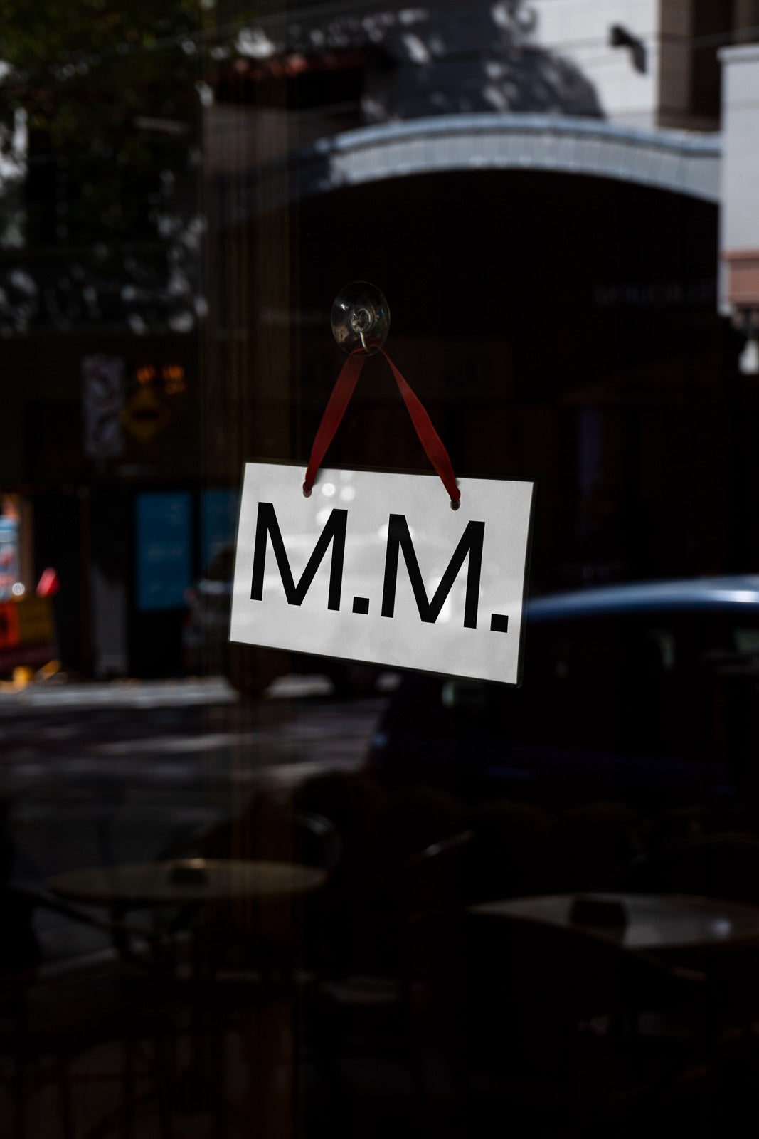 PSD mockup of a glass door with a white rectangular sign reading "M.M." hanging by a red ribbon, reflecting urban street scene in the background.