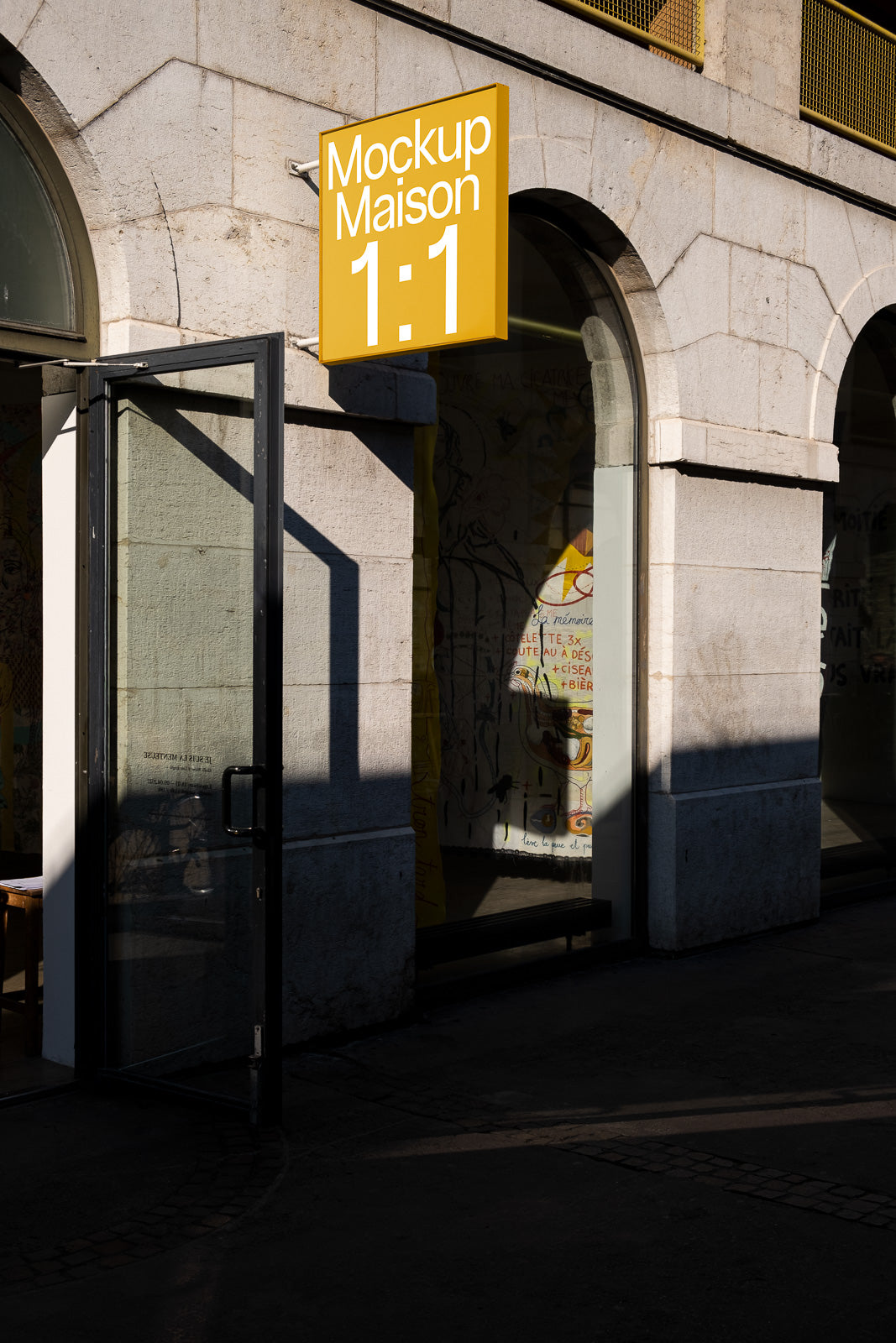 PSD mockup of a building entrance with an open glass door and a yellow sign above reading "Mockup Maison 1:1," casting shadows on the stone facade.
