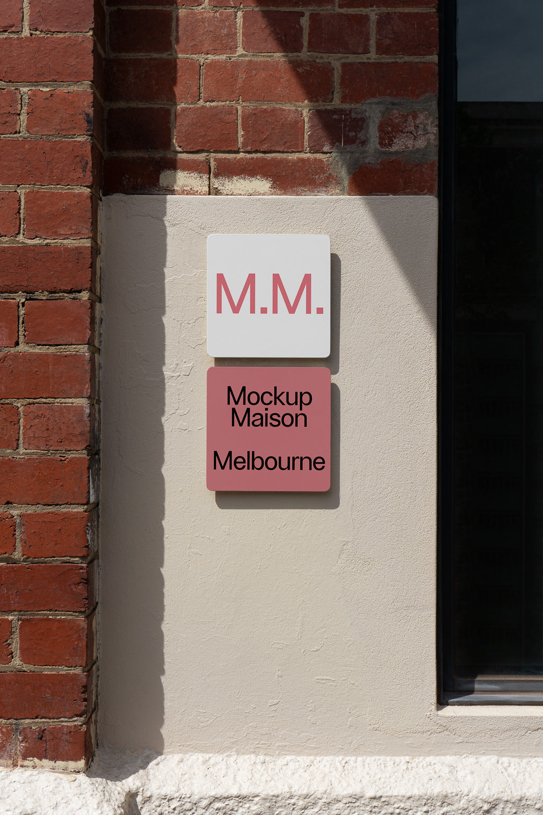 PSD mockup of a wall-mounted sign with "M.M." in pink on a white square above "Mockup Maison Melbourne" in black on a pink square against a brick wall.