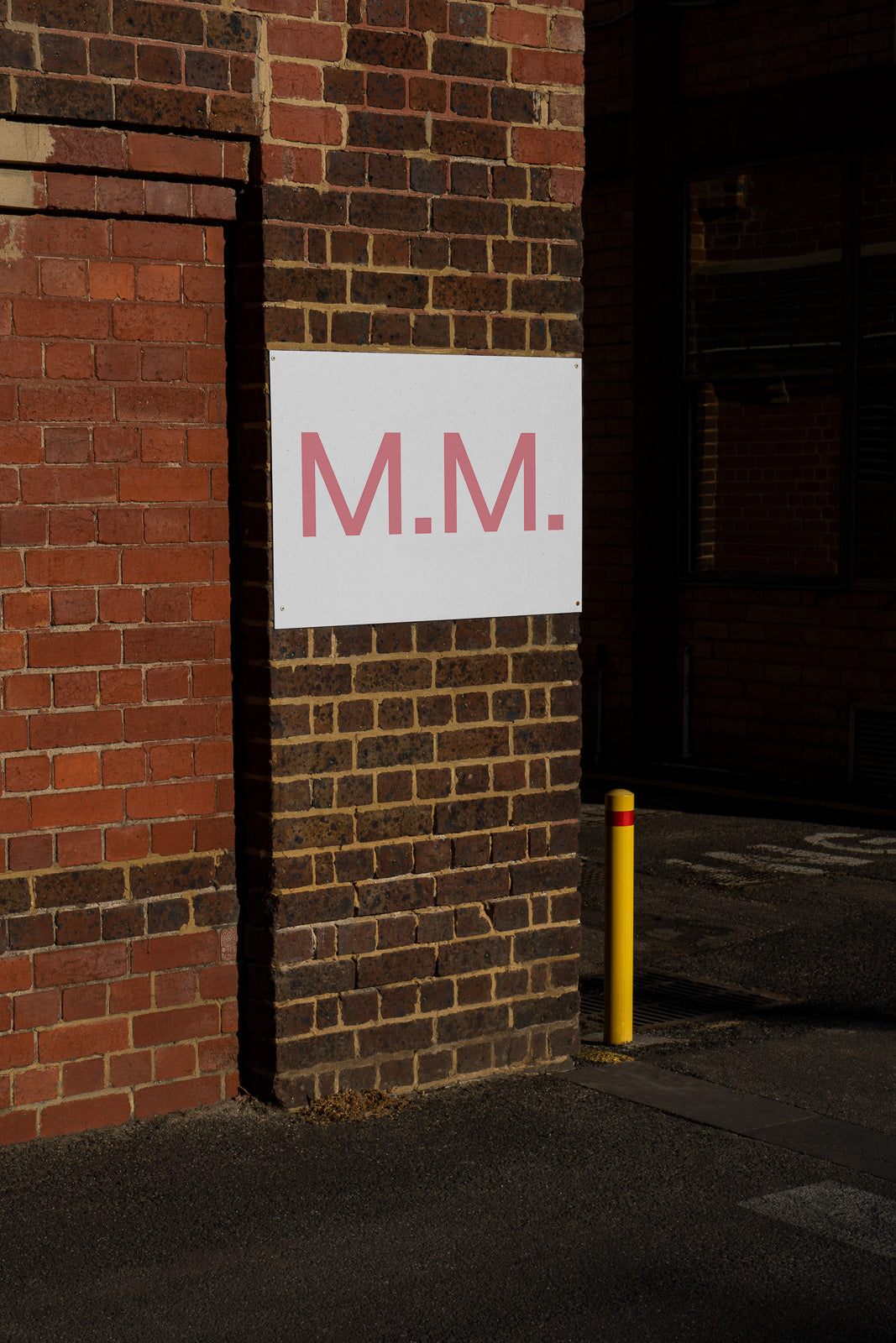PSD mockup of a sign featuring the initials "M.M." mounted on a brick wall, with a yellow bollard nearby in an urban setting.