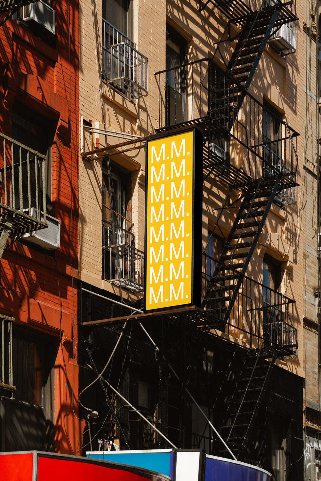 PSD mockup of a vertical building sign on a city street, featuring repeated "M.M." text in white on a yellow background, surrounded by fire escapes.