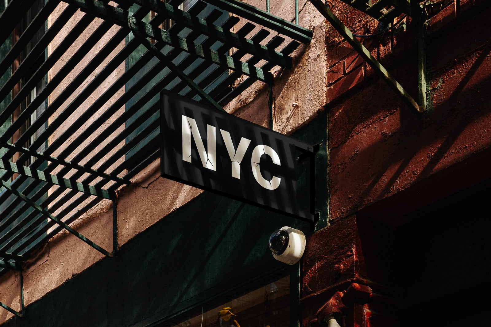 PSD mockup of a storefront sign reading "NYC" in bold white letters against a dark background, mounted on a red and green building facade with metal framework shadows.