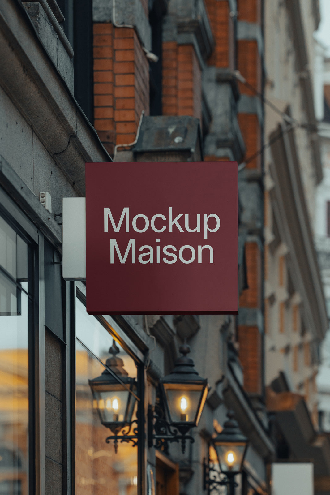 PSD mockup of a maroon rectangular storefront sign displaying "Mockup Maison" in white text, on a street with brick and stone buildings.