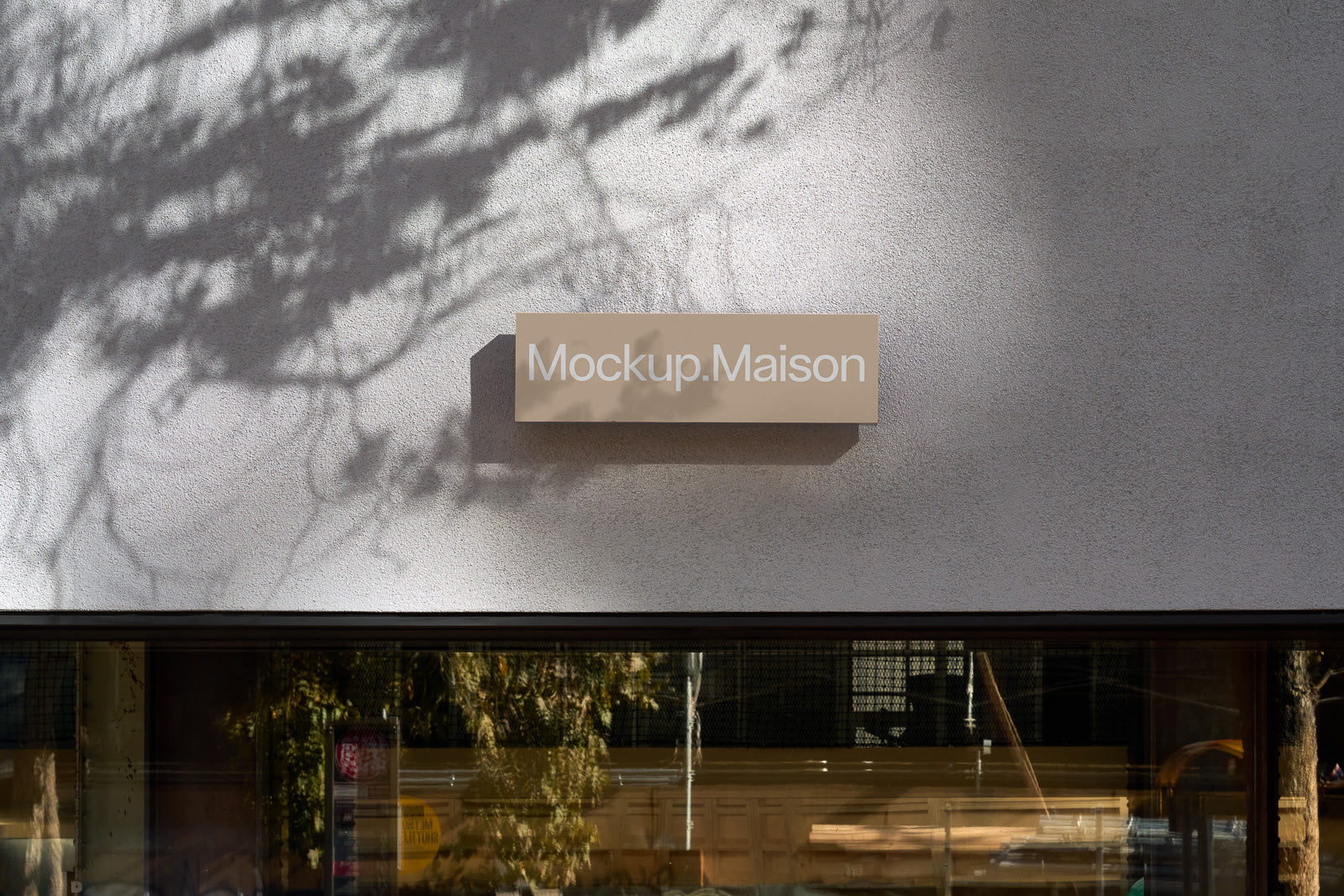 PSD mockup of a rectangular beige sign with "Mockup.Maison" text, mounted on an exterior gray wall, with tree shadows cast on the surface.