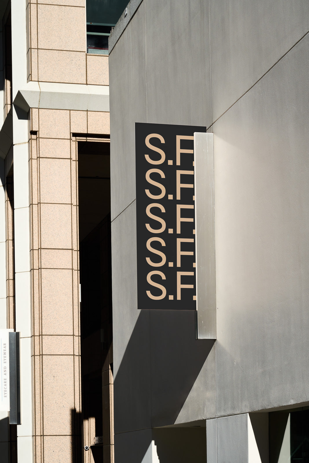 PSD mockup of a vertical building sign displaying repeated "S.F." in beige on a black background, mounted on a modern concrete wall with architectural details.