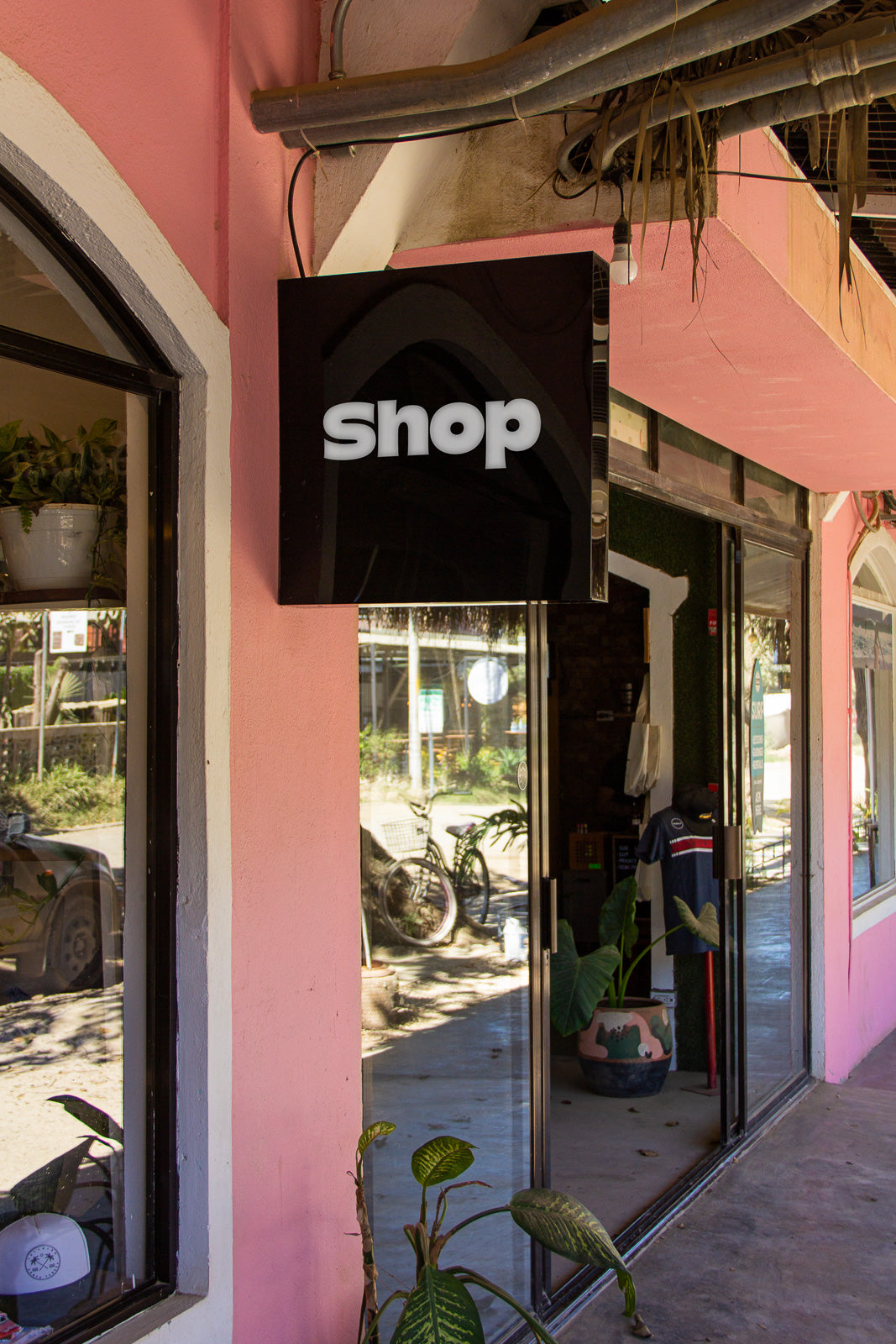 PSD mockup of a storefront with a black sign reading "shop" against a pink wall. The scene includes plants and reflections of bicycles outside the glass door.