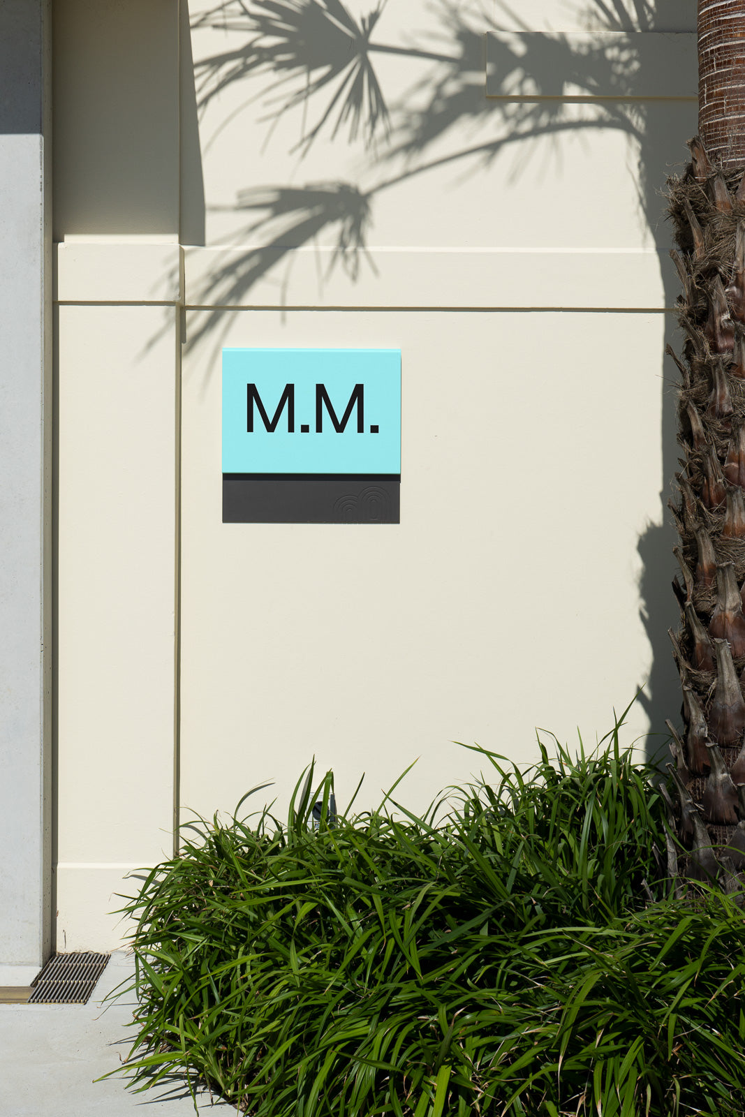 PSD mockup of an outdoor scene featuring a wall with a turquoise sign marked "M.M." in black, adjacent to a palm tree, casting shadows.