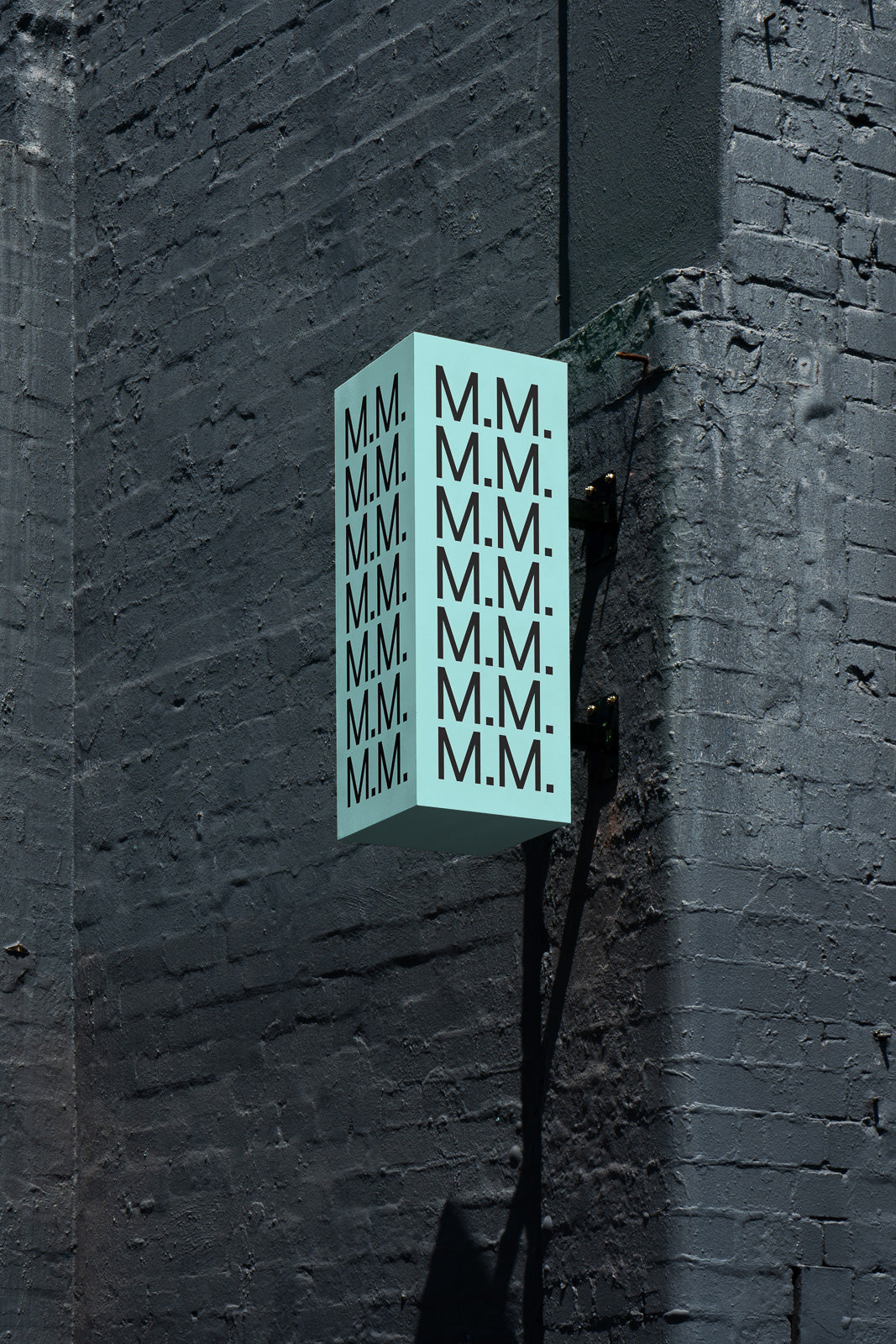 PSD mockup of a blue rectangular lightbox sign attached to a textured dark brick wall, featuring repeated black "M.M." text on its side.