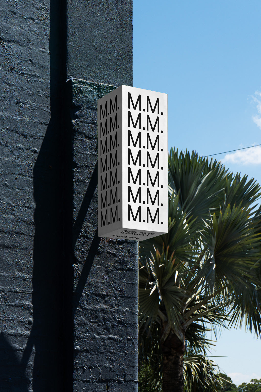 PSD mockup of a rectangular hanging sign on a dark brick wall, featuring repeated "M.M." text, with palm trees in the background against a clear sky.
