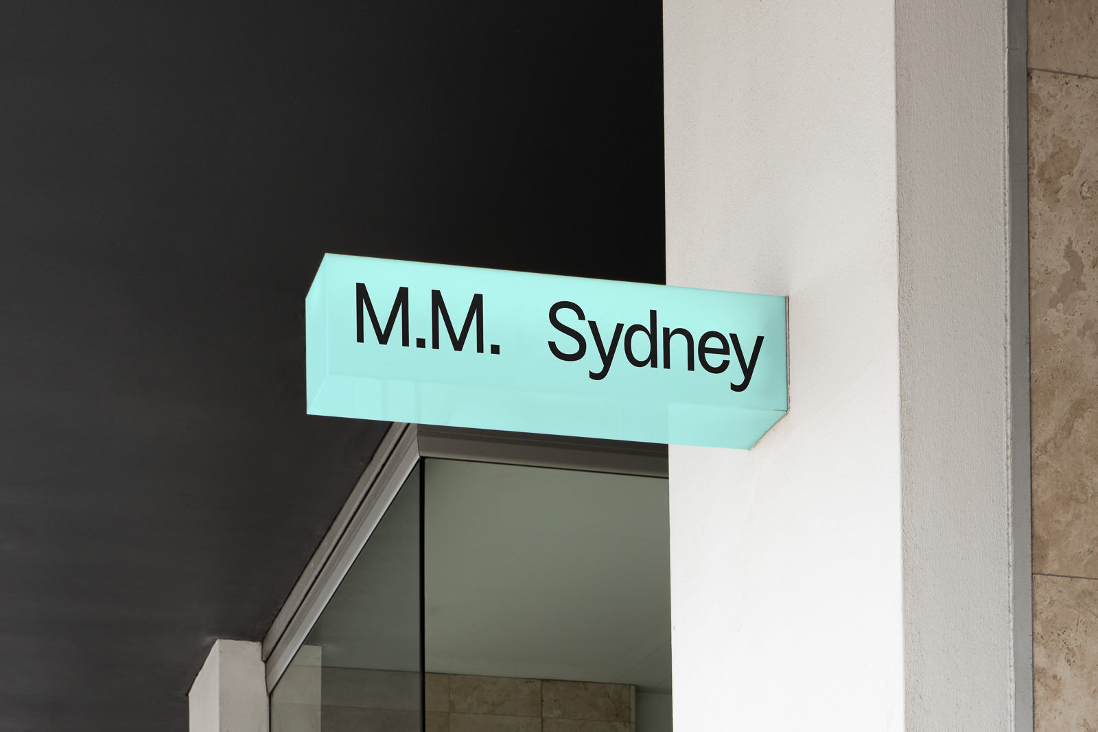 PSD mockup of a turquoise rectangular sign with "M.M. Sydney" in bold black letters attached to a building column, set against a modern facade.