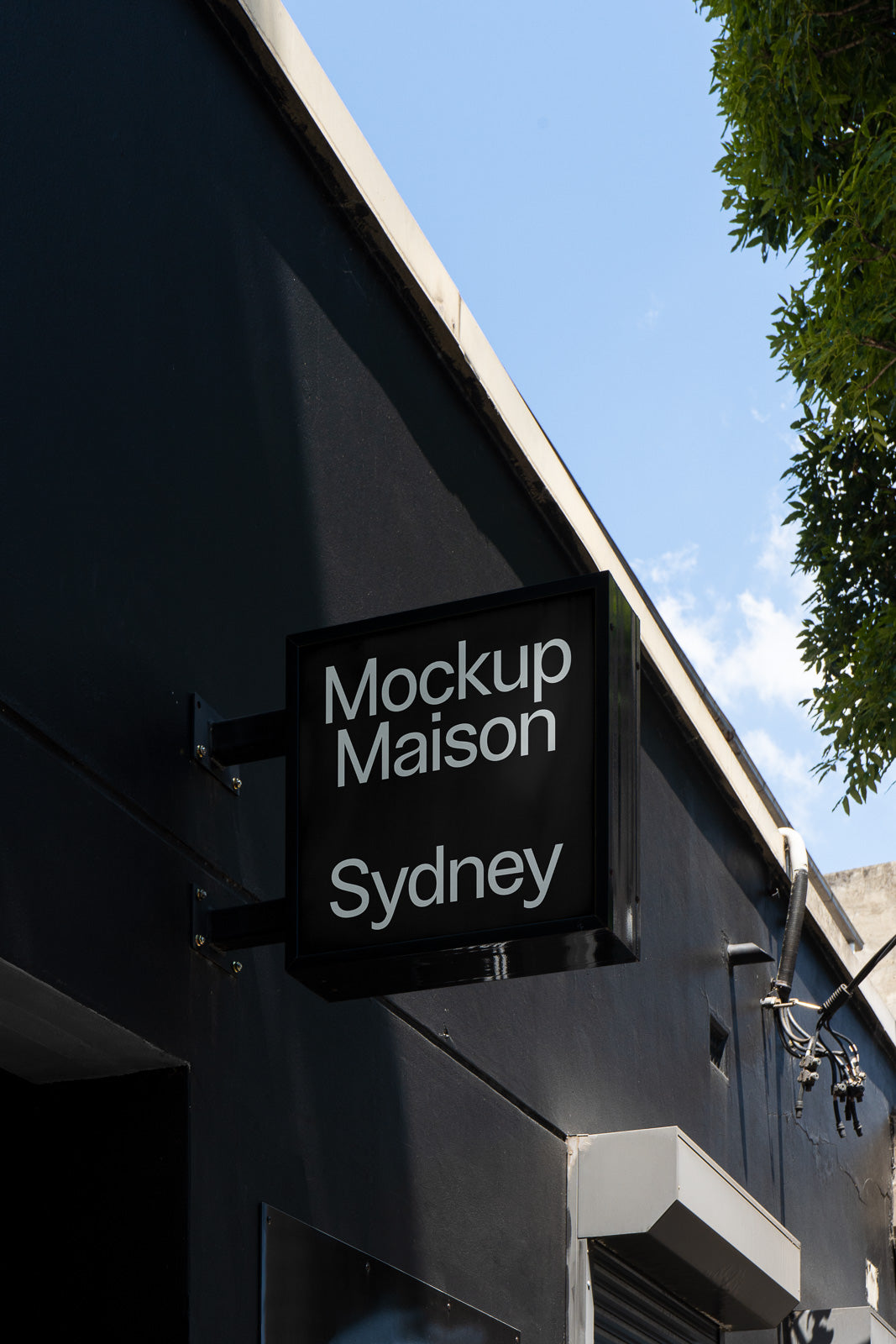 PSD mockup of a rectangular store sign attached to a dark building facade, with "Mockup Maison Sydney" written in white against the blue sky.