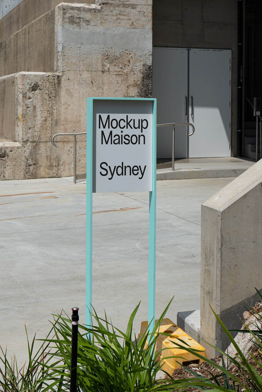 PSD mockup of an outdoor sign with a turquoise frame reading "Mockup Maison Sydney" against a textured concrete wall near stairs.