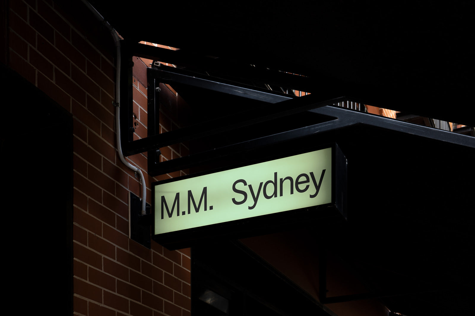 PSD mockup of an illuminated sign displaying "M.M. Sydney" mounted on a brick wall under a dark awning.