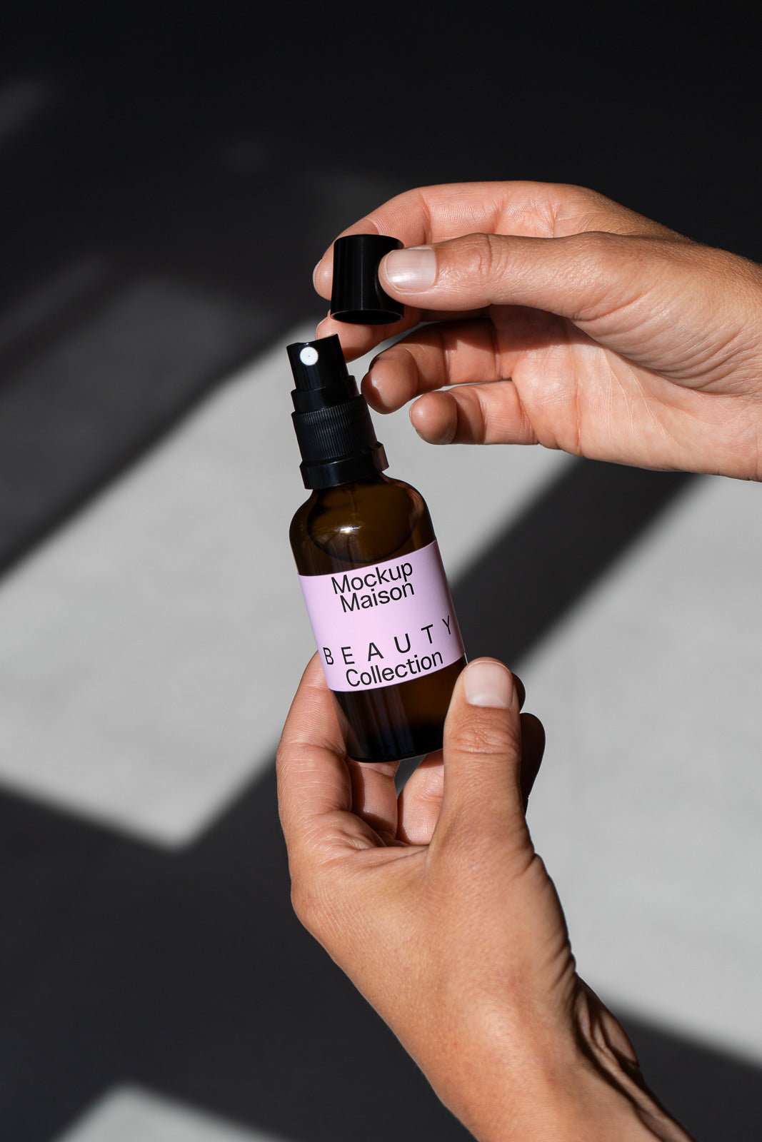 PSD mockup of a hand holding a spray bottle labeled "Mockup Maison Beauty Collection" against a shadowed background with sunlight.