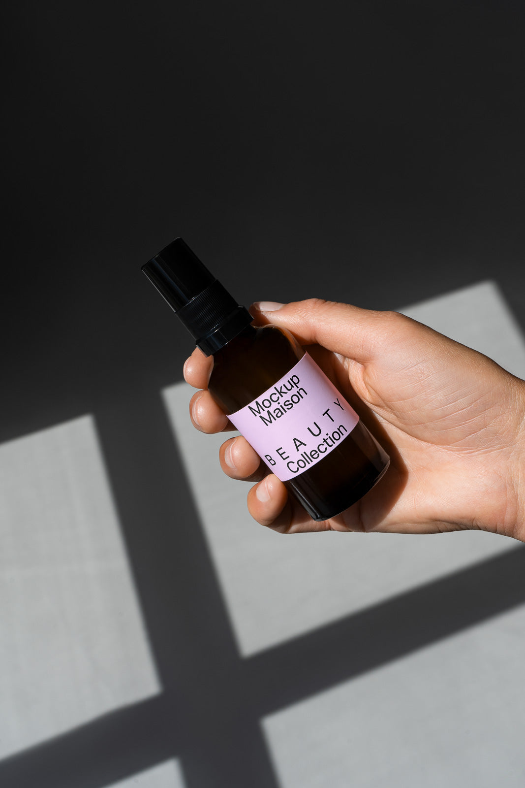PSD mockup of a hand holding a black spray bottle labeled "Mockup Maison Beauty Collection" against a gray background.