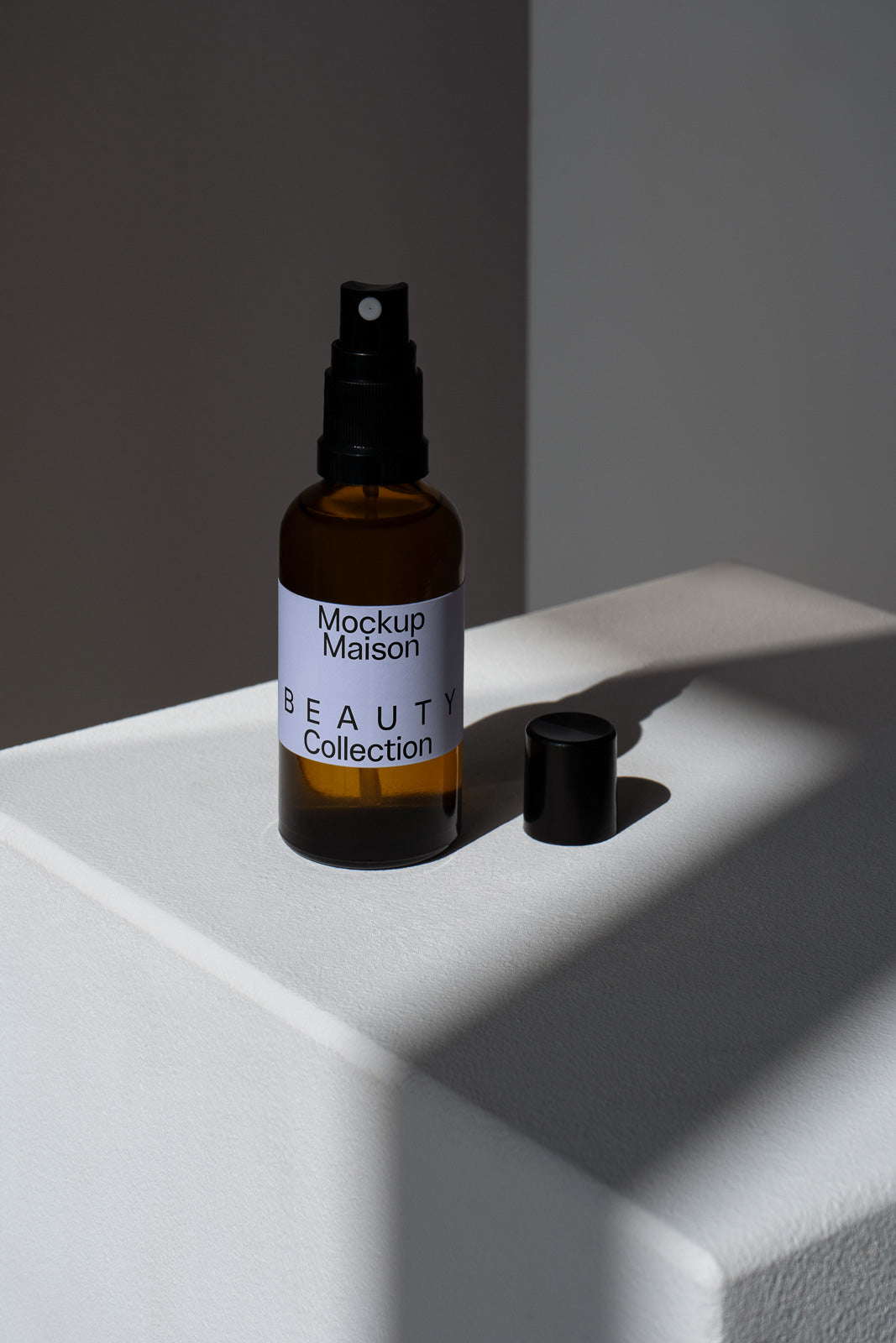 PSD mockup of a brown spray bottle labeled "Mockup Maison Beauty Collection" on a white surface with a detached black cap nearby in soft lighting.