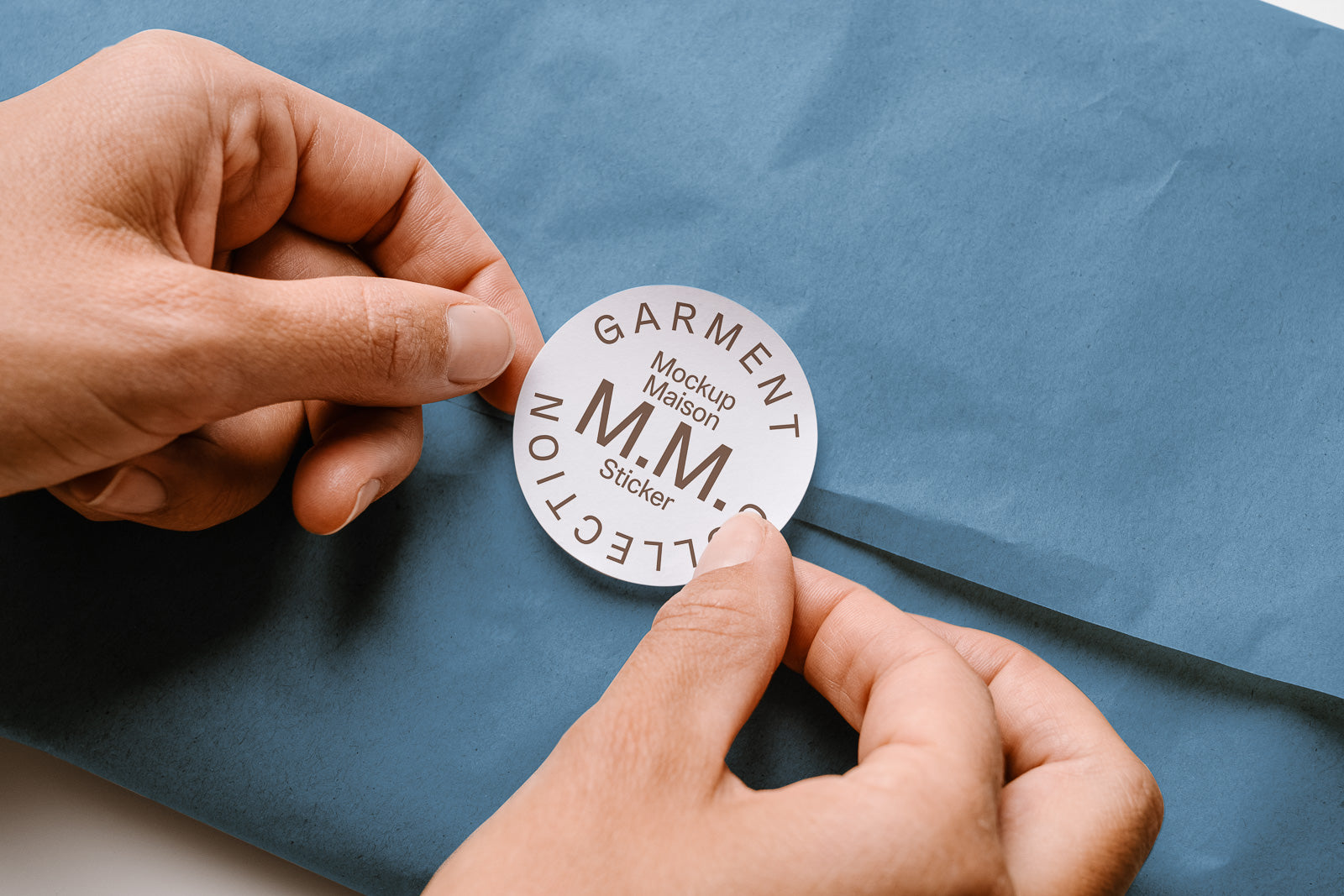 PSD mockup of hands applying a round sticker labeled "Garment Collection, Mockup Maison M.M. Sticker" onto blue packaging paper.