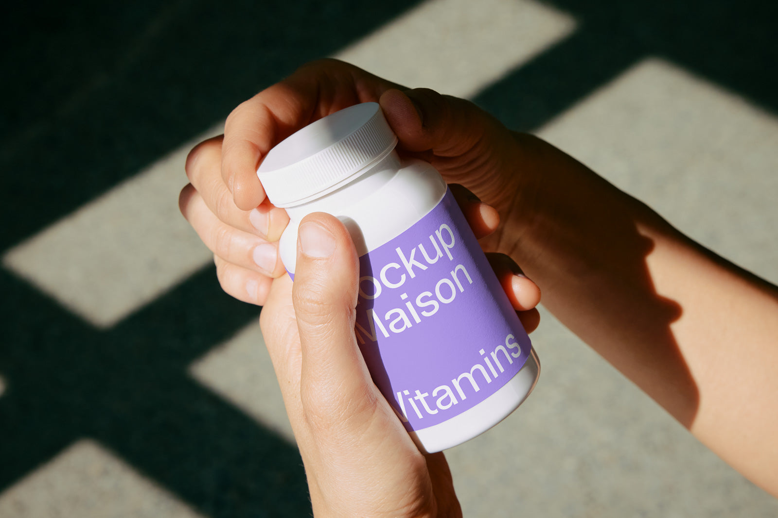 PSD mockup of a person holding a white bottle with a purple label featuring text. The lid is being opened, and the background is softly lit with patterned shadows.
