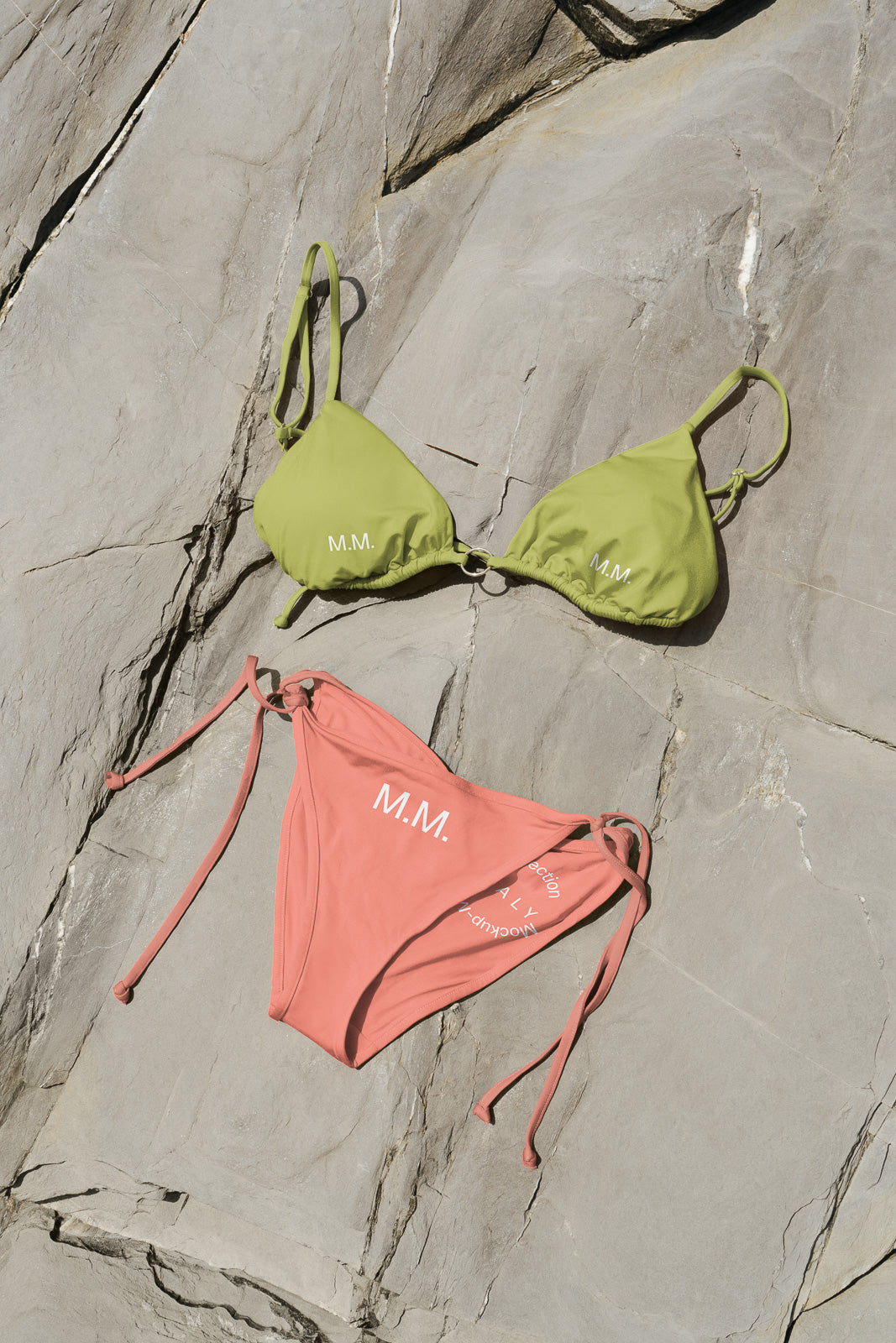 PSD mockup of a two-piece bikini set on textured stone background, featuring olive green top and coral bottom with "M.M." initials.
