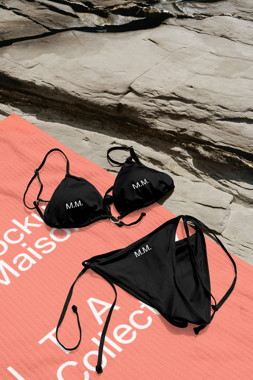 PSD mockup of a black bikini with "M.M." initials on a coral beach towel, set against a rocky background.