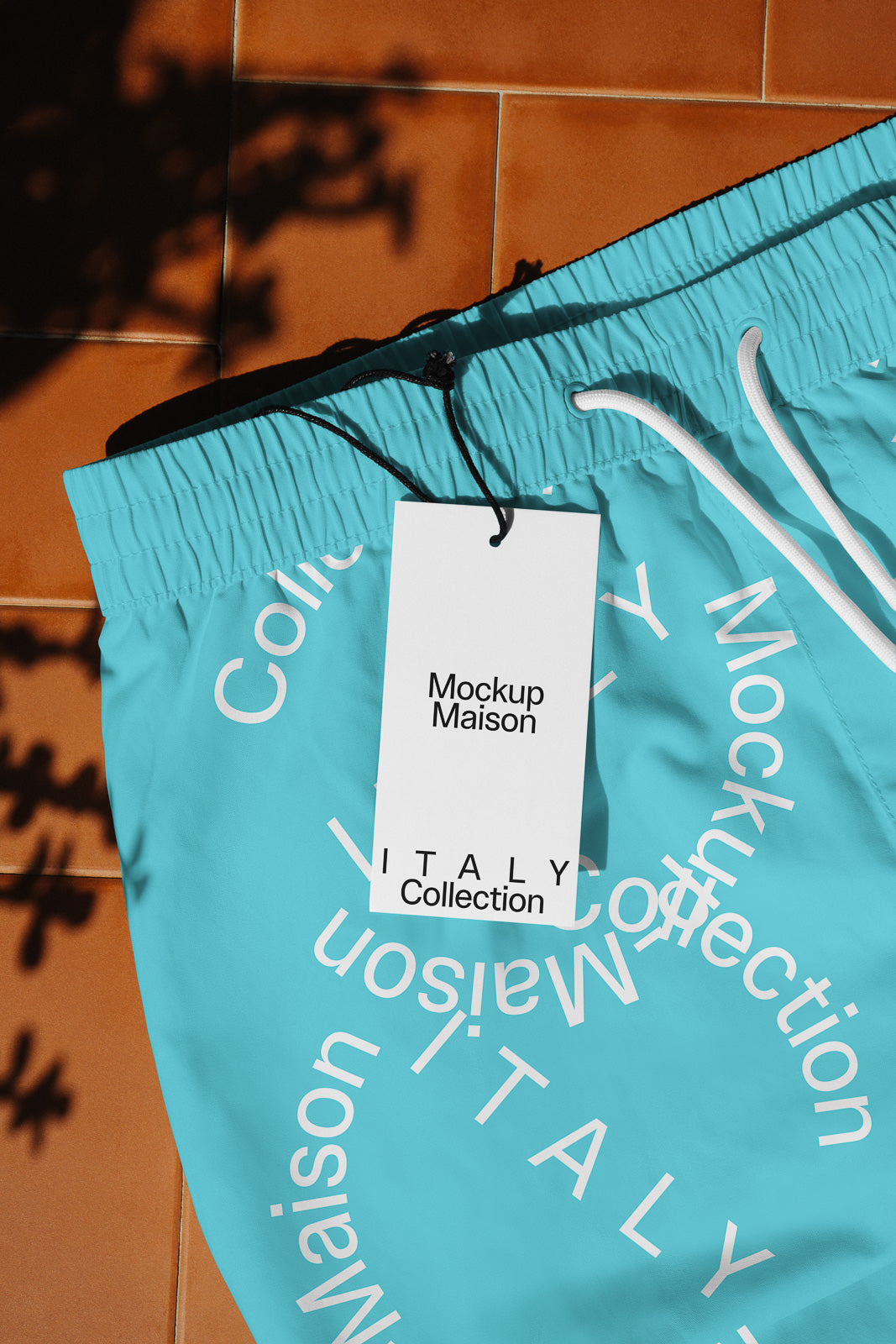 PSD mockup of turquoise shorts with white drawstrings, featuring a tag labeled "Mockup Maison" and "Italy Collection" on a terracotta tile background.