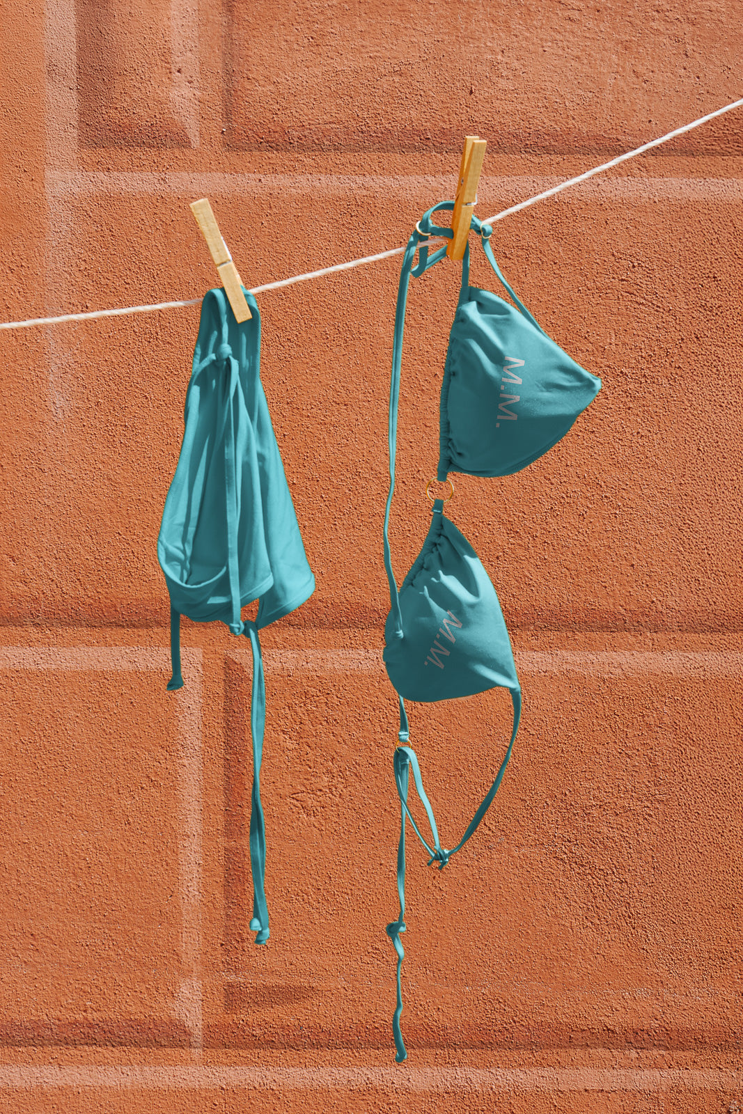 PSD mockup of turquoise bikini with top and bottom pieces hanging on a clothesline against a textured orange wall background.