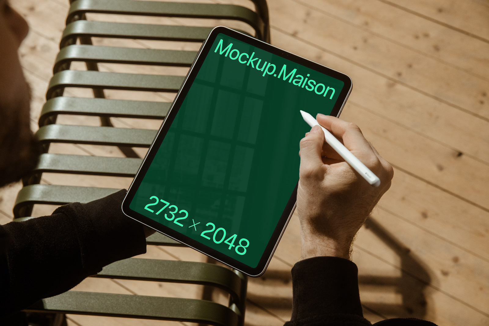 PSD mockup of a person using a stylus on a tablet with a green screen displaying "Mockup.Maison" and dimensions "2732 x 2048" on a bench with a wooden floor background.