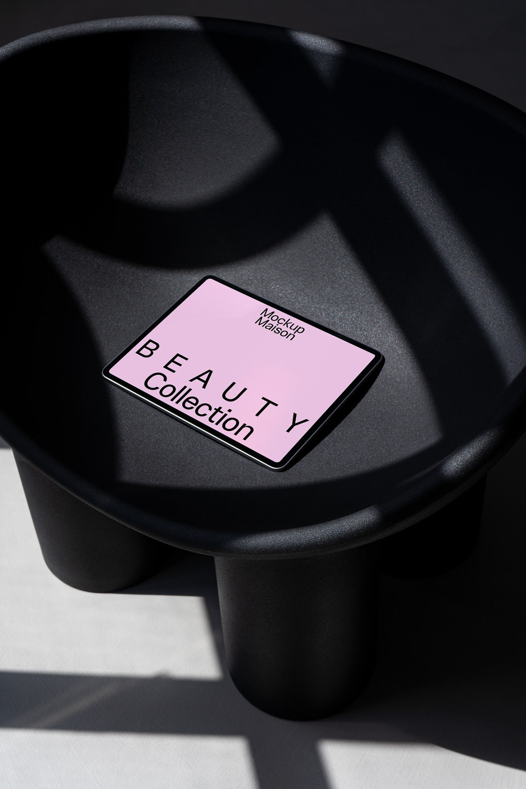 PSD mockup of a tablet displaying "Beauty Collection" text in a stylized font on a pink screen, placed on a modern dark chair with shadow patterns.