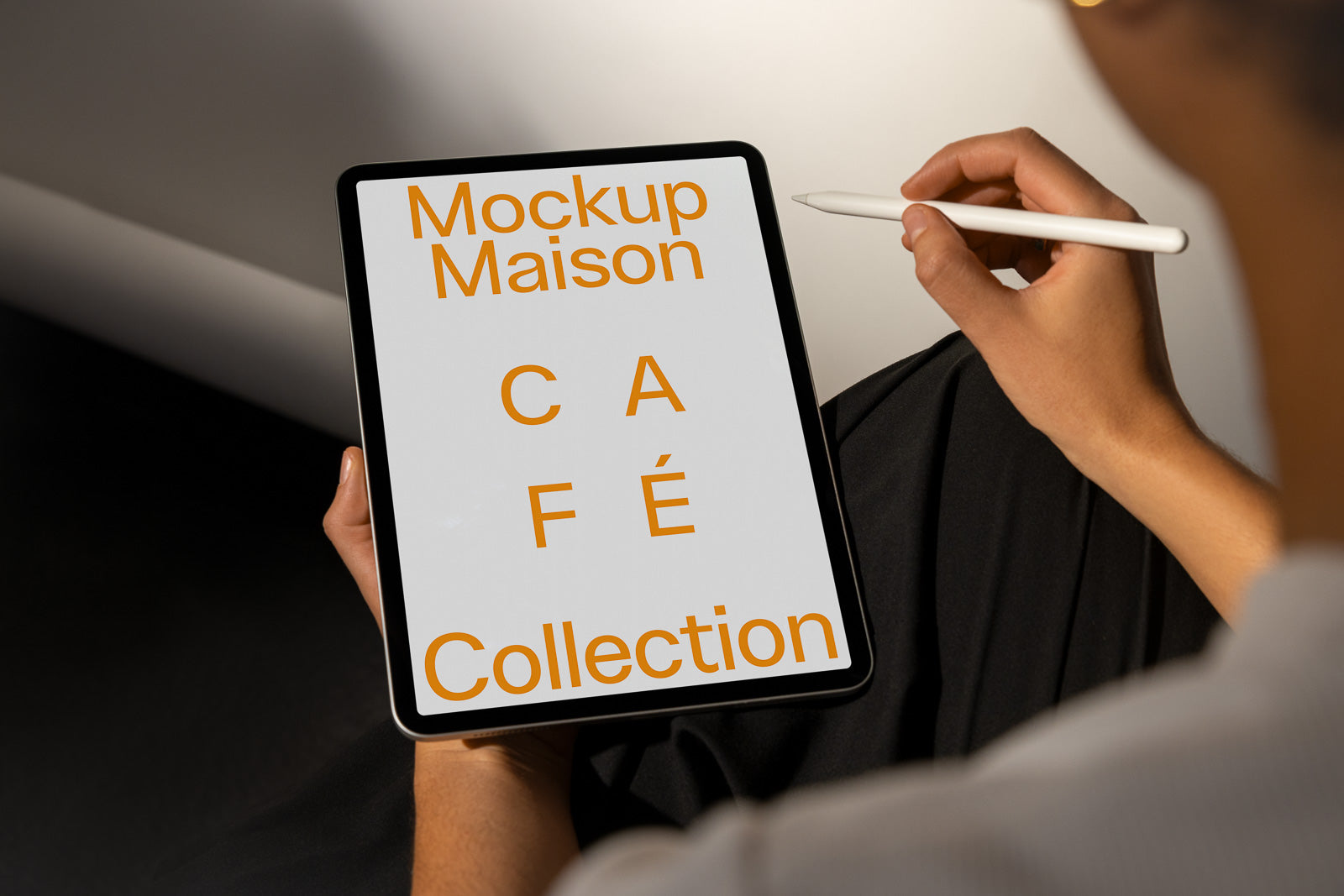 PSD mockup of a person holding a tablet displaying text that reads "Mockup Maison Café Collection" in orange font, with a stylus in hand.