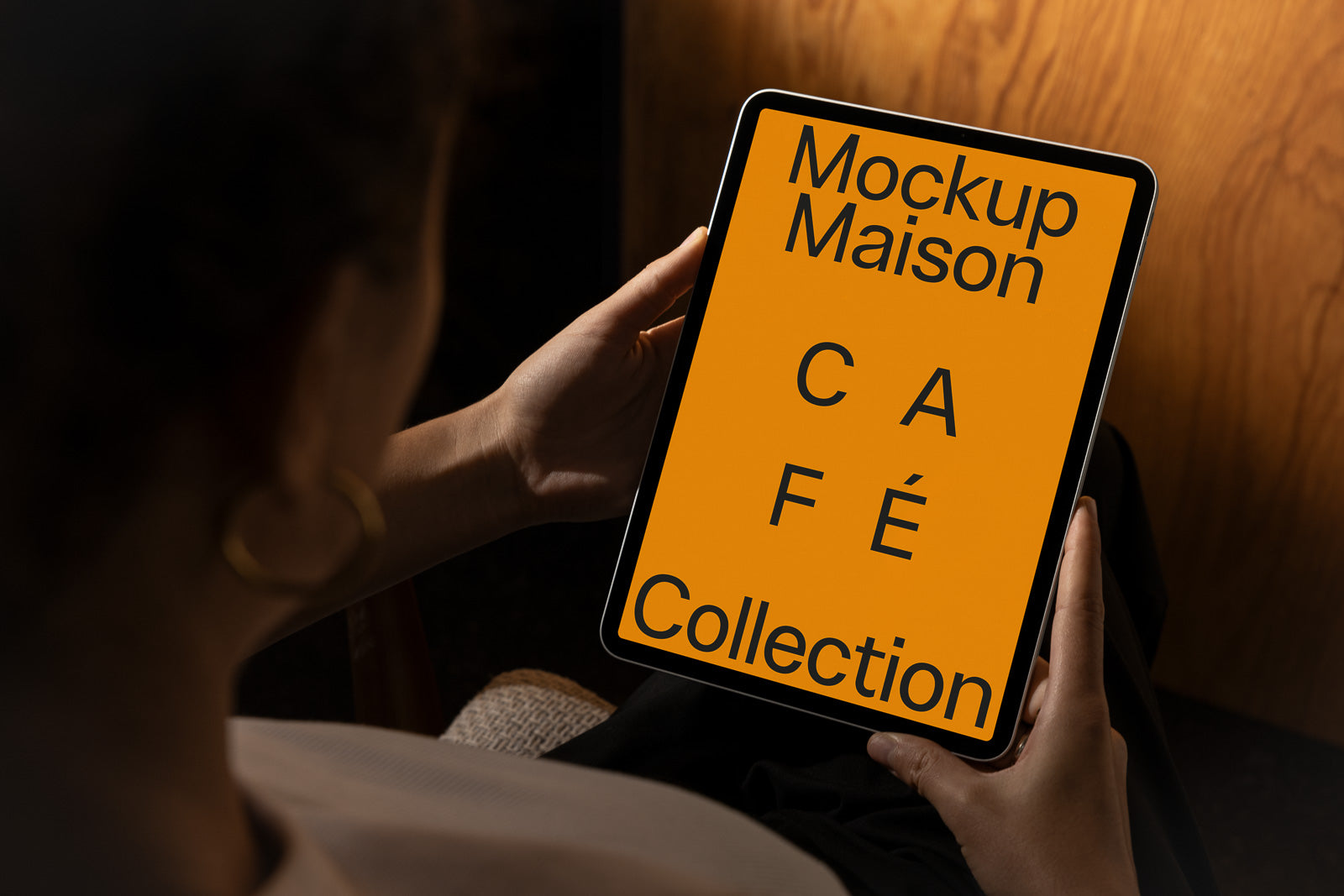 PSD mockup of a person holding a tablet displaying "Mockup Maison Café Collection" on a bright orange screen, seated in a dimly lit room with wood-paneled background.