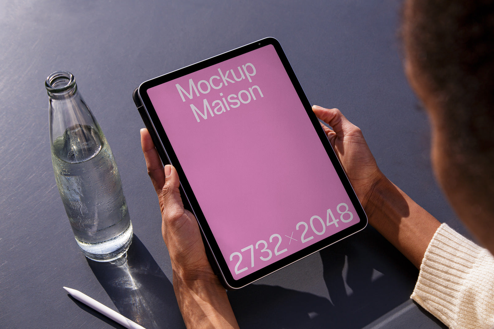 PSD mockup of a tablet held in hands with a pink screen displaying text "Mockup Maison" and "2732 x 2048," next to a bottle and stylus on a table.