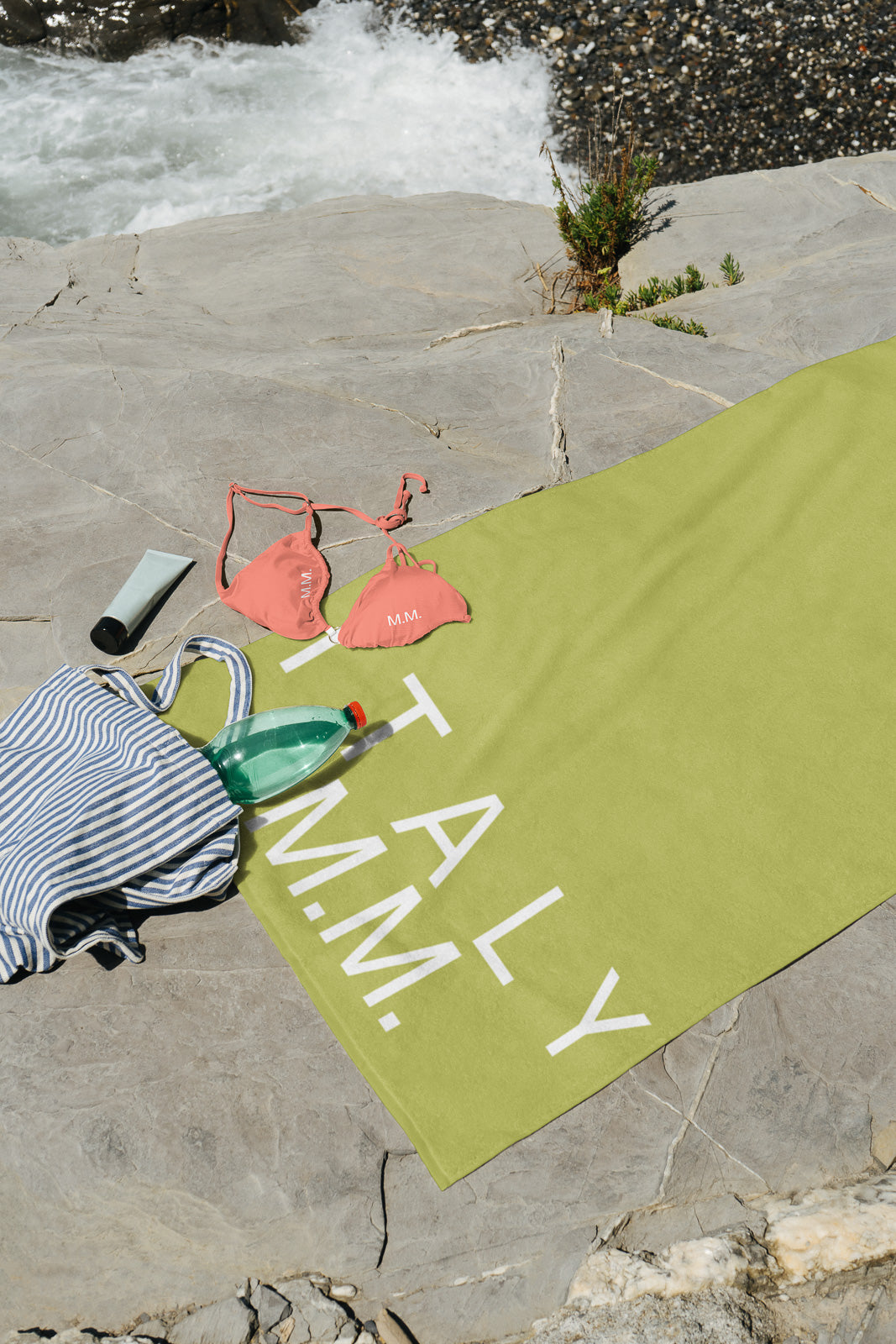 PSD mockup of a beach scene with a green towel on rocks, a striped bag, bottled water, sunscreen, and coral bikini top near the ocean.