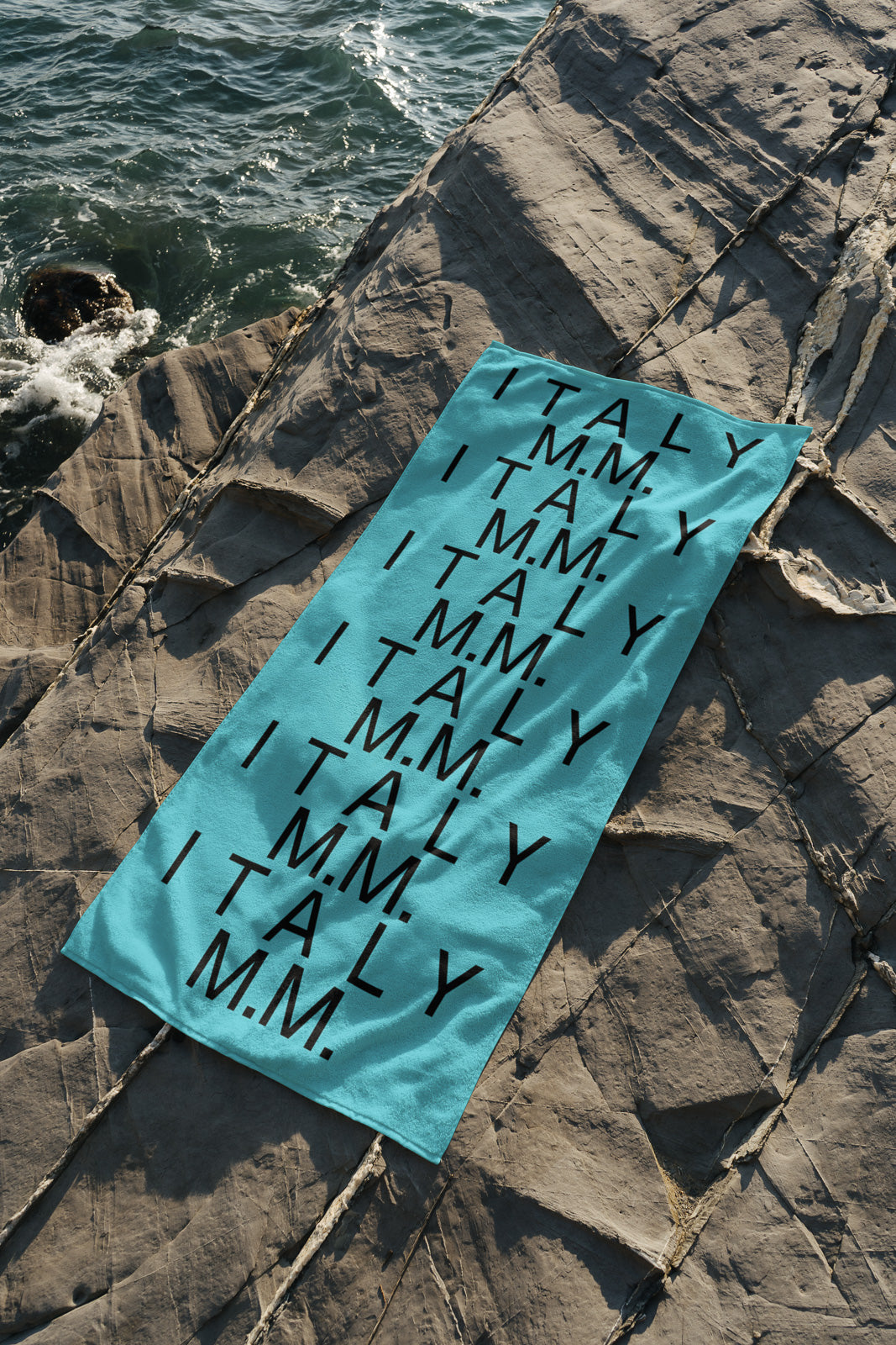 PSD mockup of a turquoise towel with repeated black text "ITALY M.M." placed on rocky terrain near the ocean.