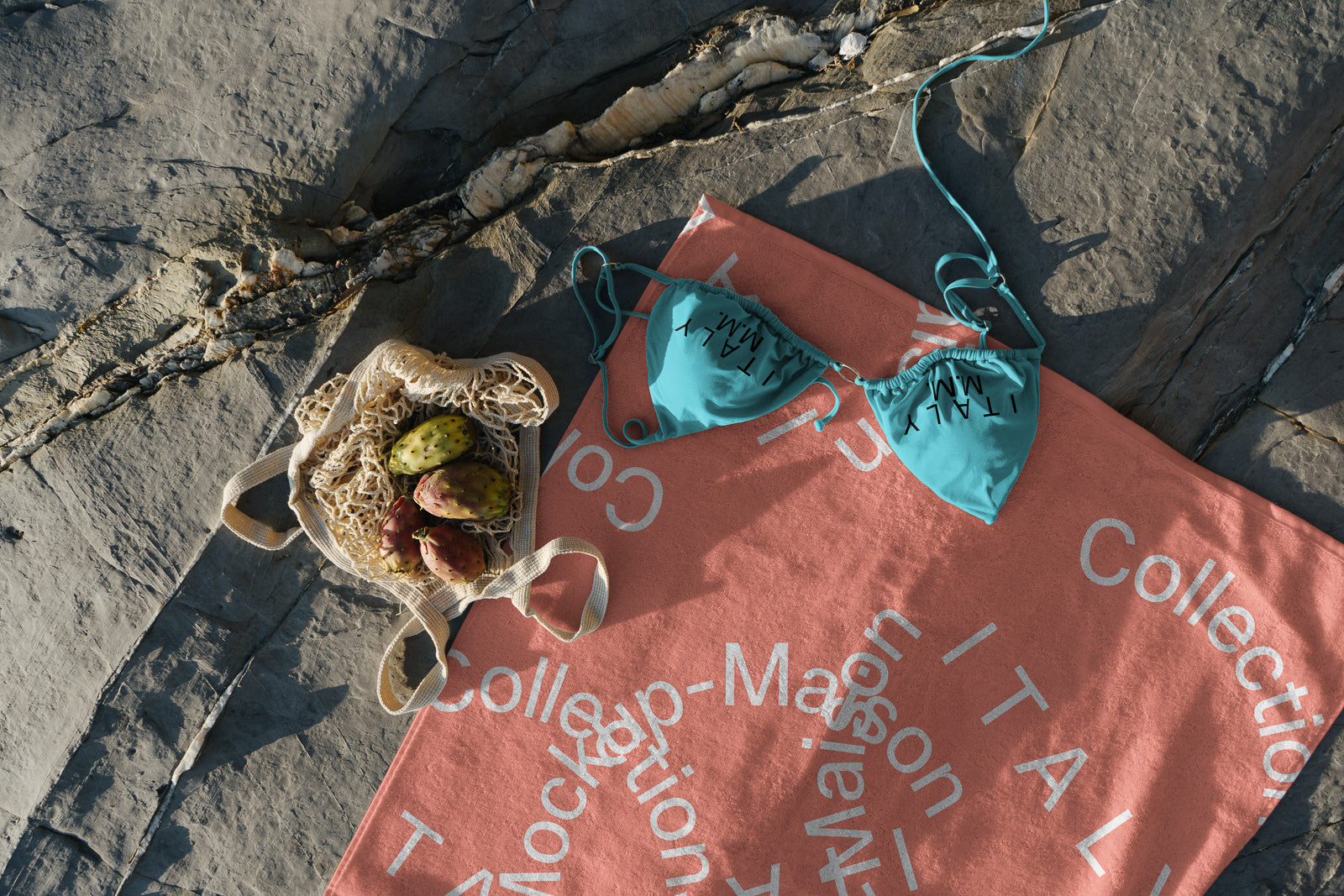 PSD mockup of a teal bikini on a coral towel with abstract text, laid on rocks beside a string bag containing prickly pears.