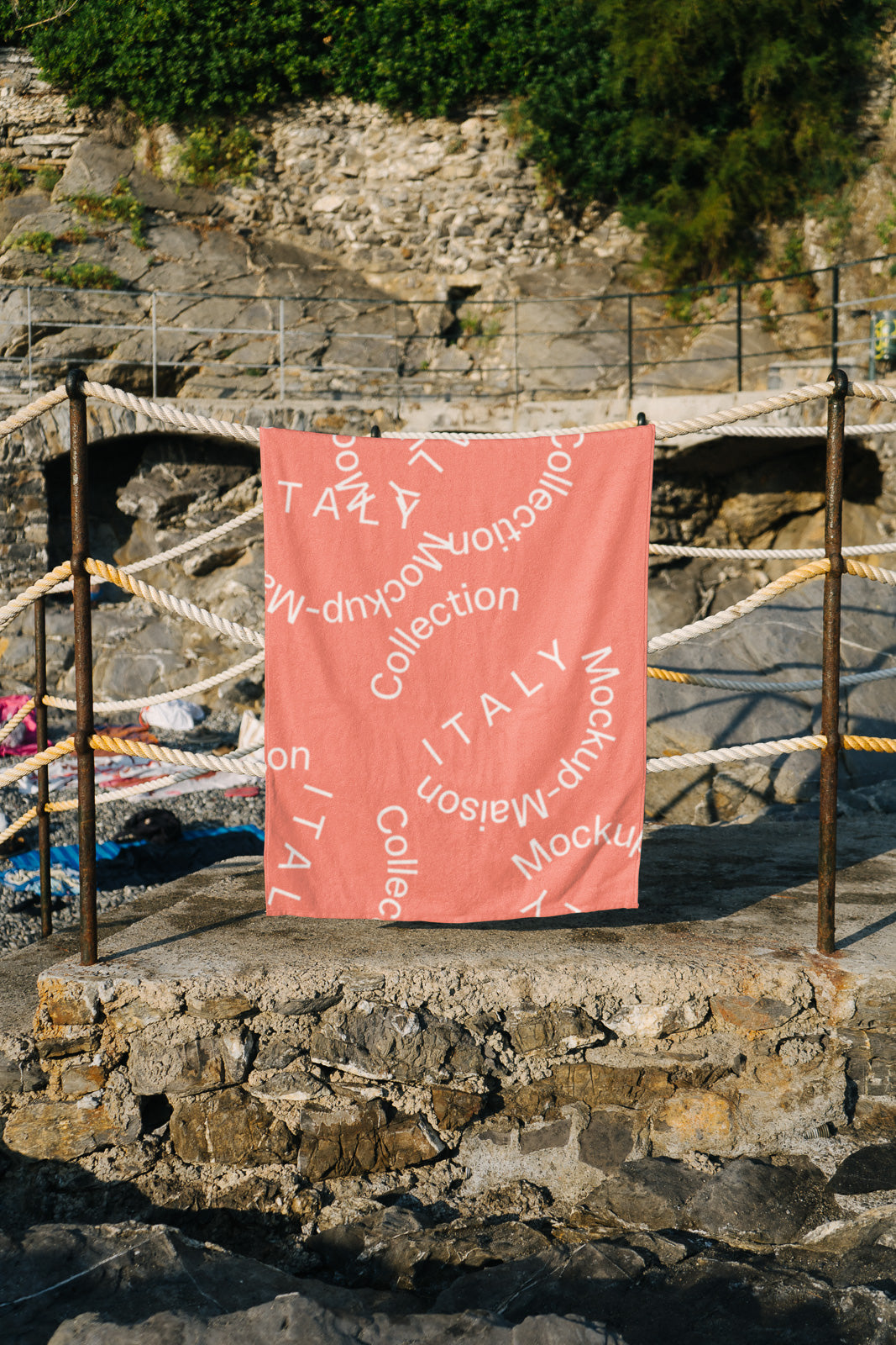 PSD mockup of a pink towel with white swirling text saying "ITALY Collection" hanging on a rope fence, set against a rocky outdoor backdrop.