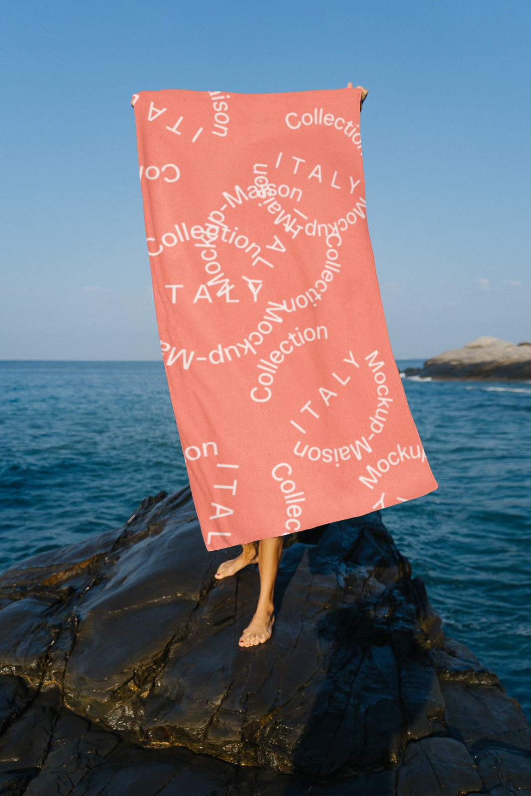 PSD mockup of a pink towel with text patterns, held by a person standing on rocks by the sea under a clear blue sky.