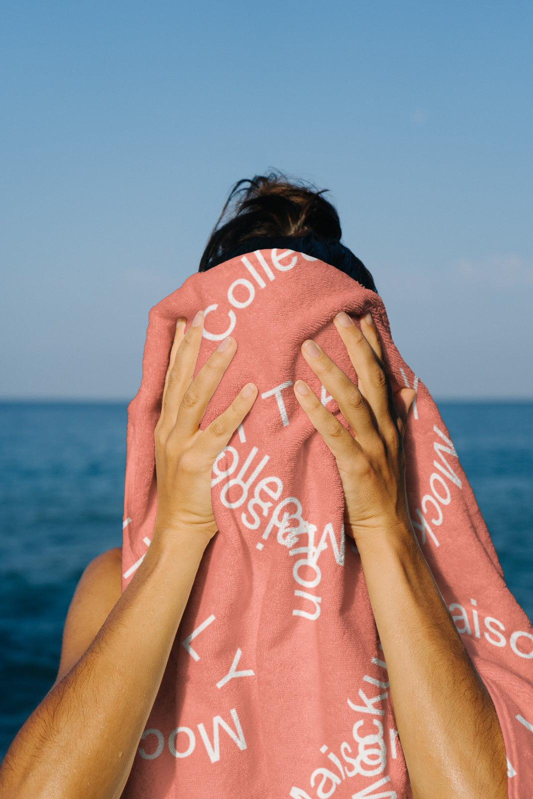 PSD mockup of a person holding a coral towel with white text against a clear blue sky and ocean backdrop, partially covering their face.