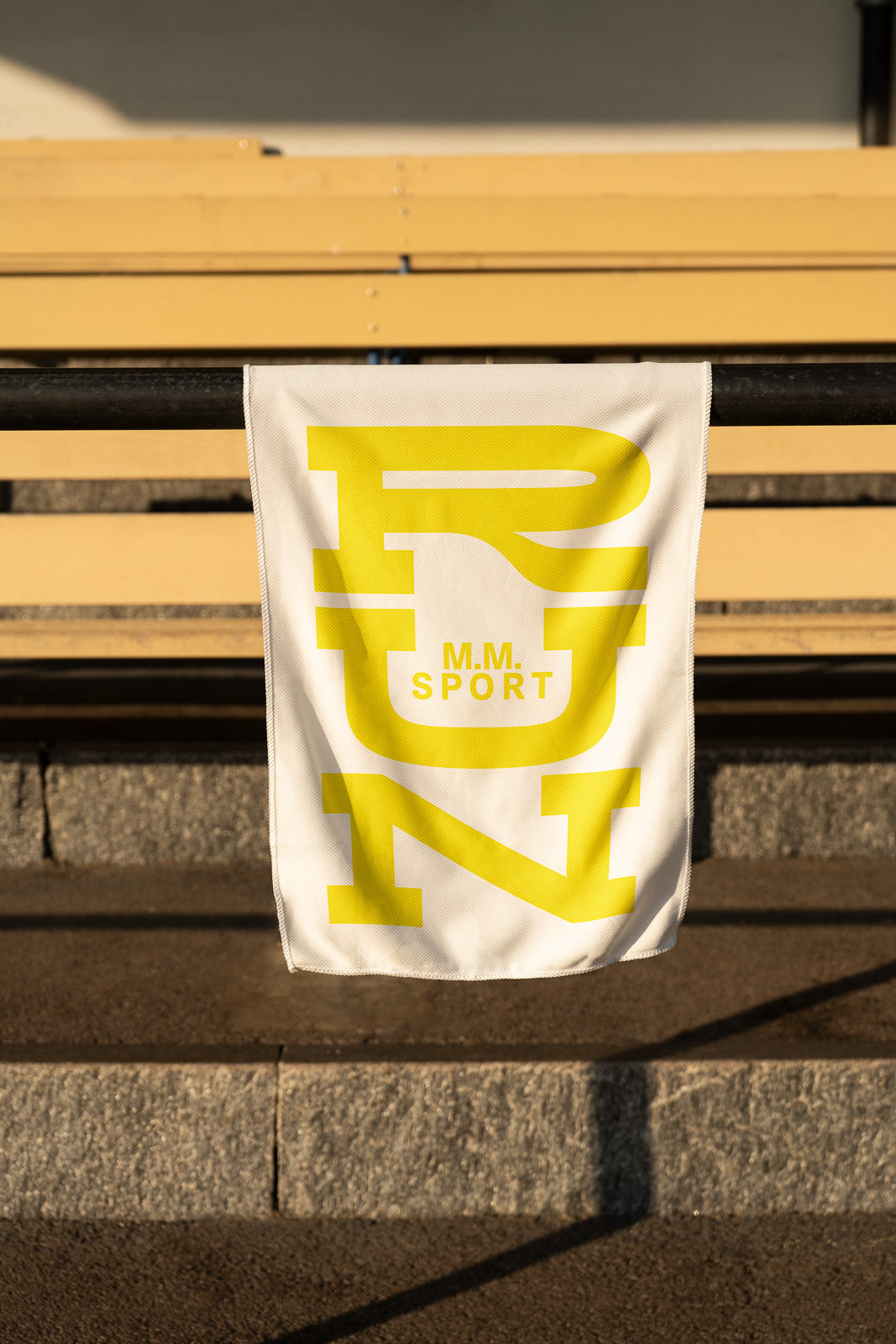 PSD mockup of a white banner draped over a black metal rail with a yellow "RUN" design and "M.M. SPORT" text, set against a wooden bench and stone steps.