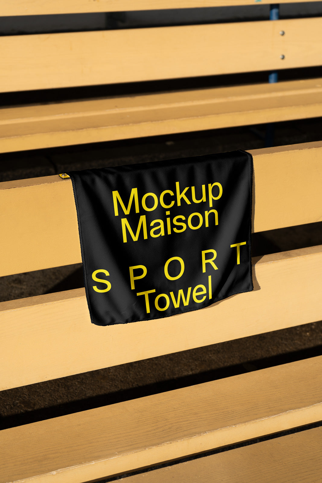 PSD mockup of a black sports towel with yellow text "Mockup Maison SPORT Towel," draped over a wooden bench in an outdoor setting.