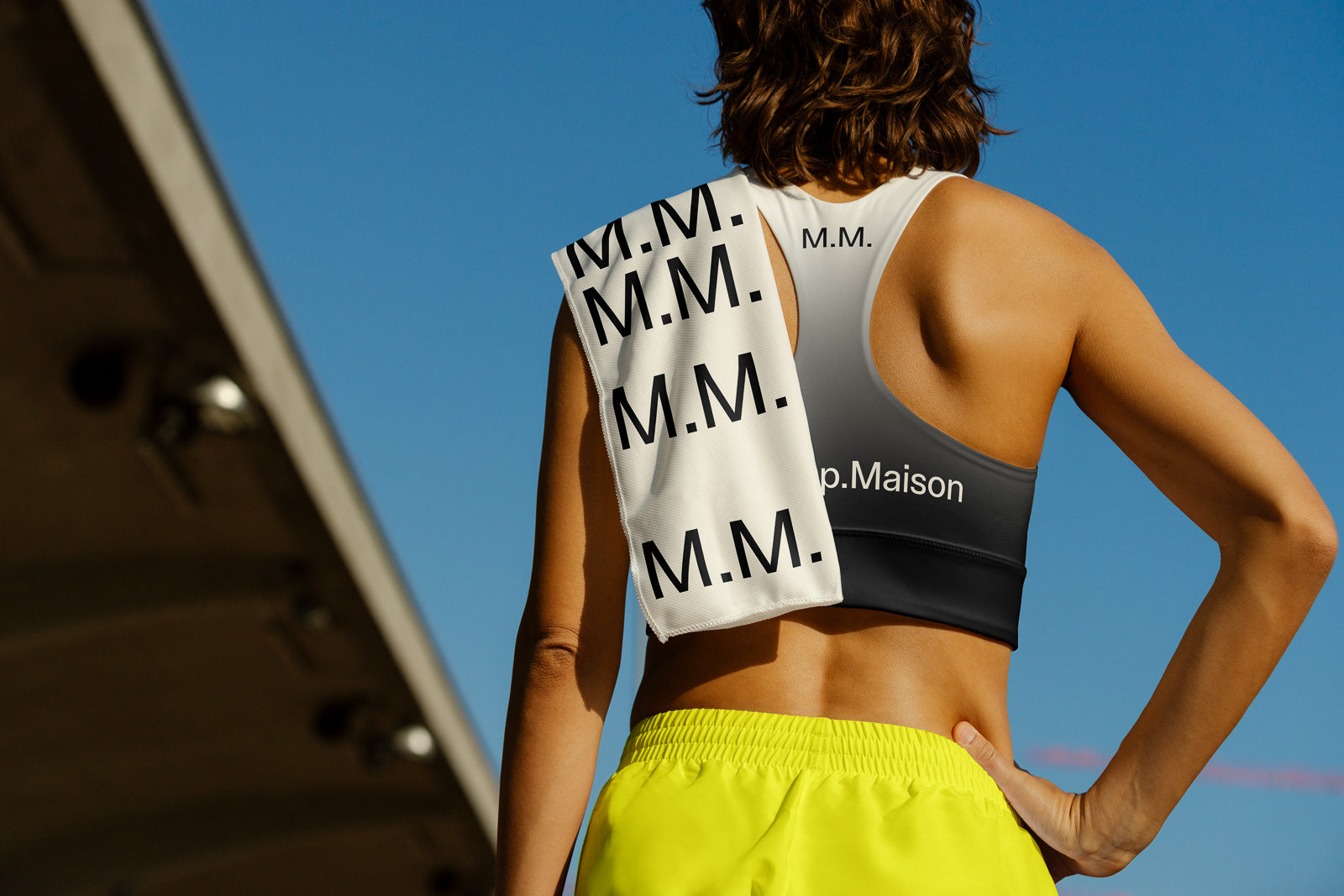 PSD mockup of a person wearing a gradient sports bra with "M.M." text, holding a matching towel over the shoulder, bright shorts, clear blue sky.