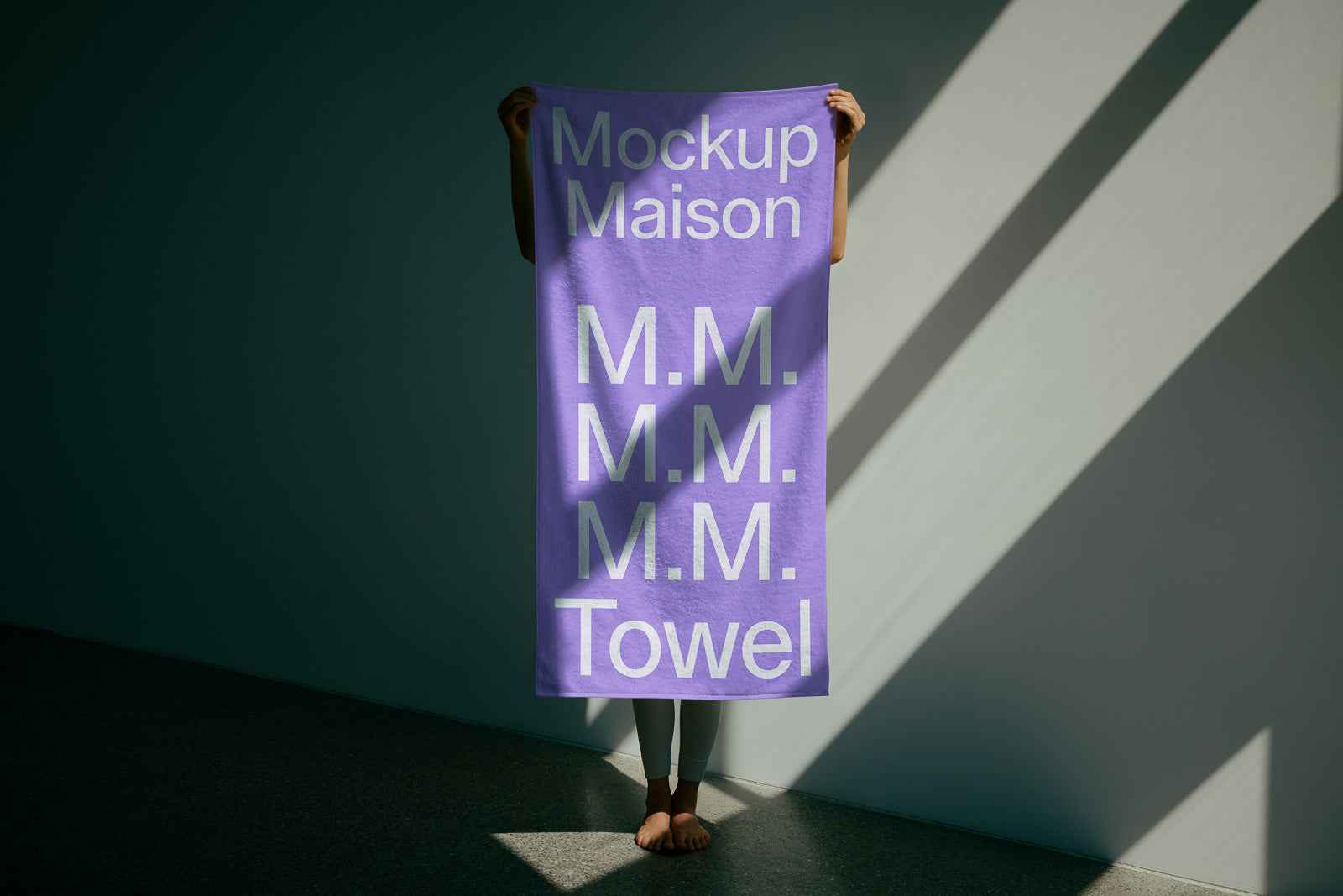PSD mockup of a purple towel held vertically, featuring white text "Mockup Maison" and "M.M." repeated with "Towel" at the bottom. Background in shadow.