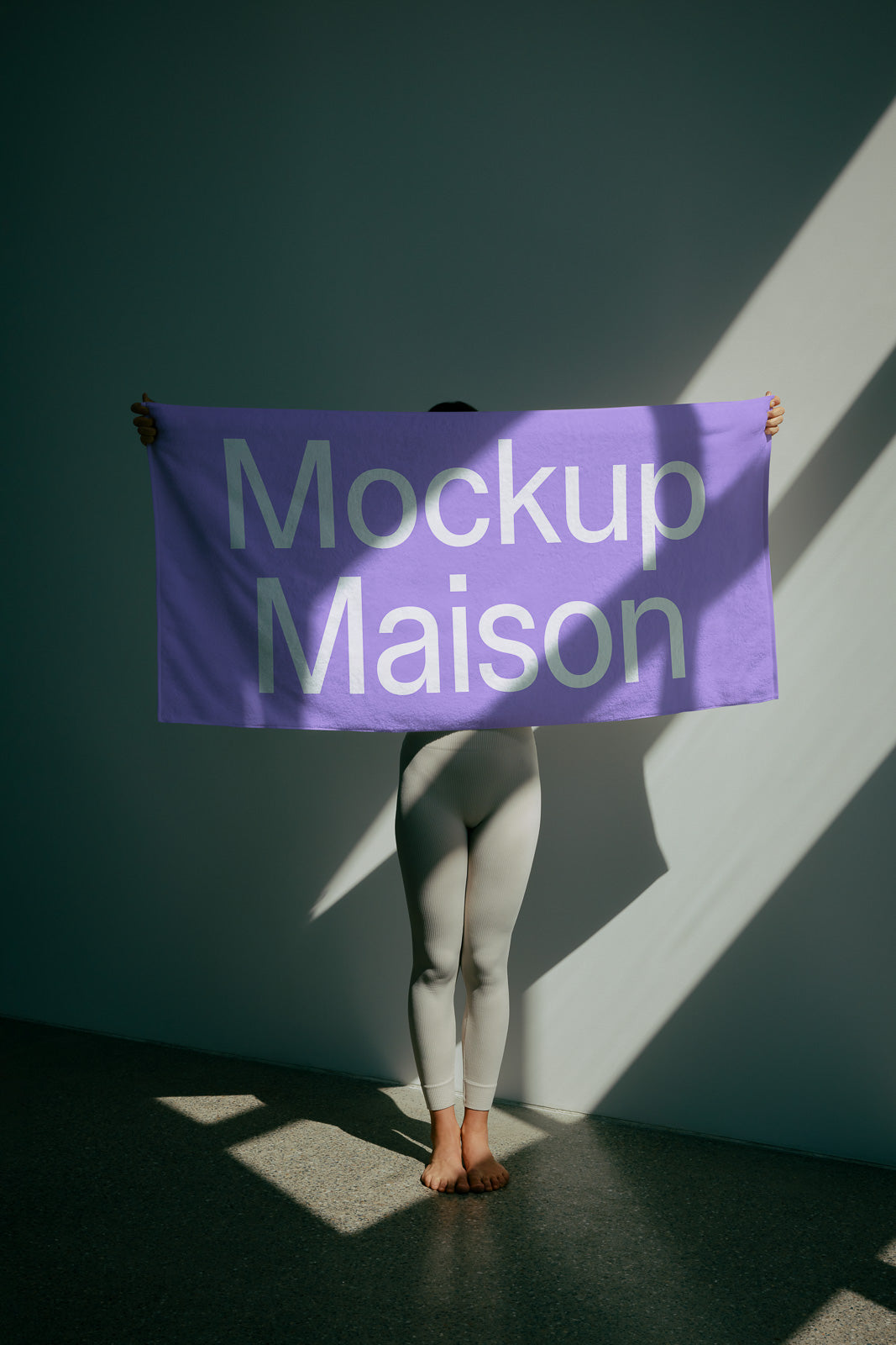 PSD mockup of a purple towel being held in front of a person, casting shadows on a wall in a sunlit room.