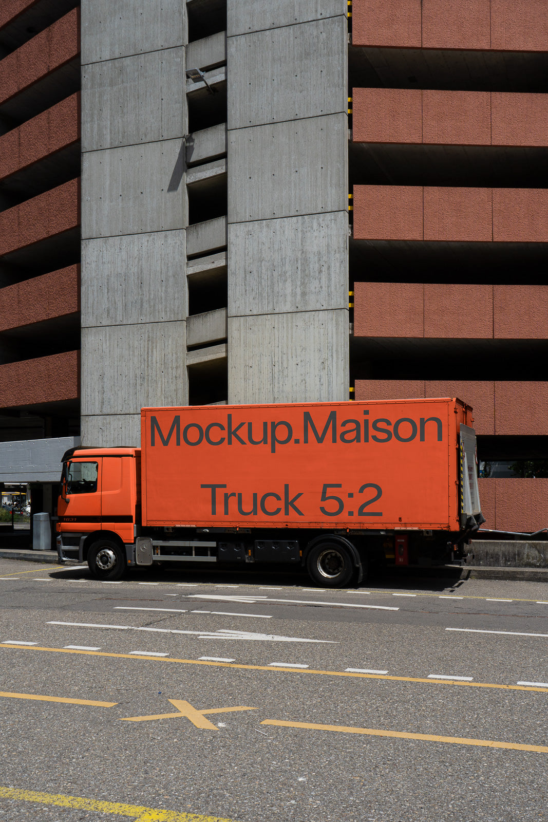 PSD mockup of an orange truck parked beside a concrete and brick building, displaying "Mockup.Maison Truck 5:2" in bold letters on its side.
