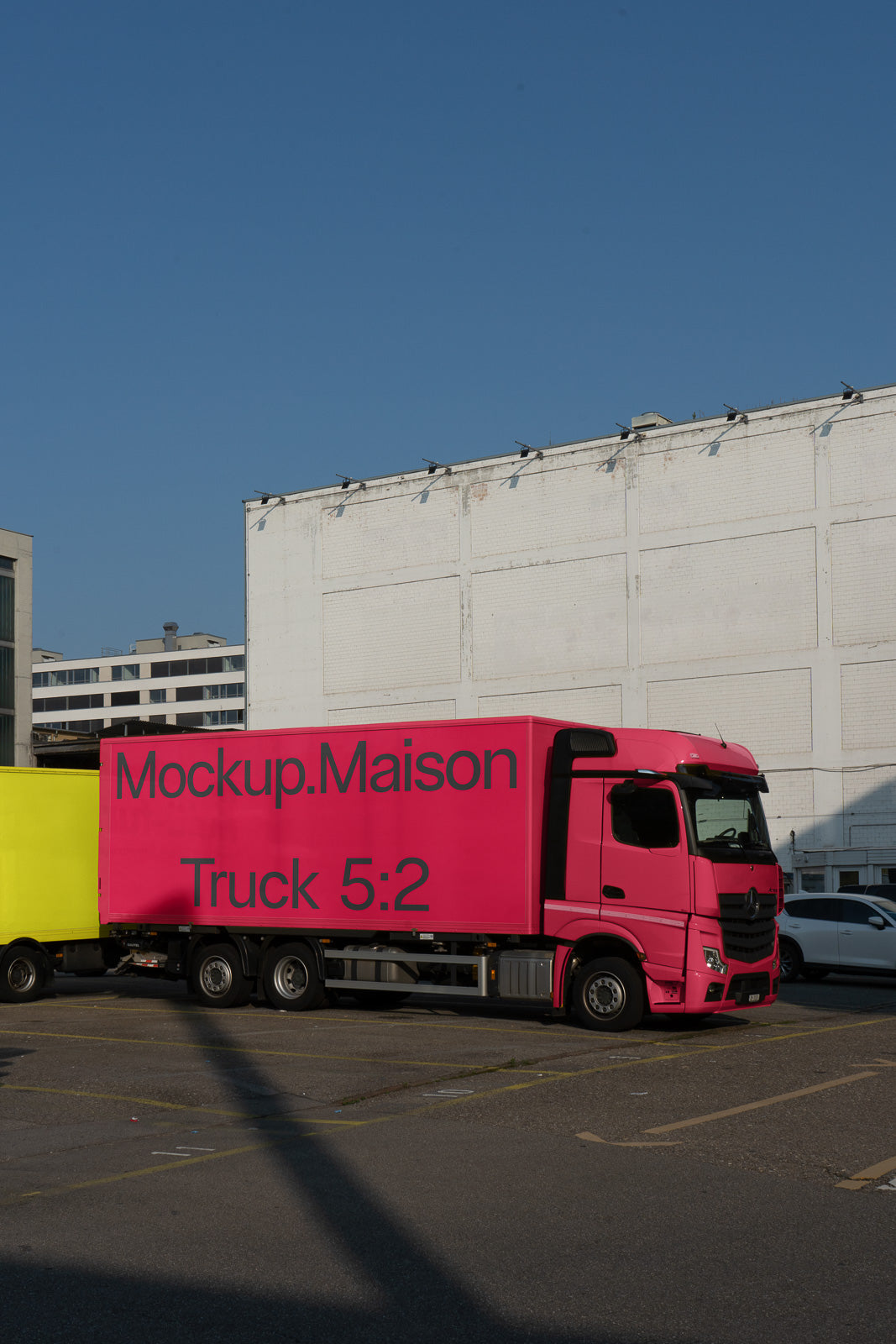 PSD mockup of a bright pink truck labeled "Mockup.Maison Truck 5:2," parked next to a yellow truck against a plain white building under a clear blue sky.
