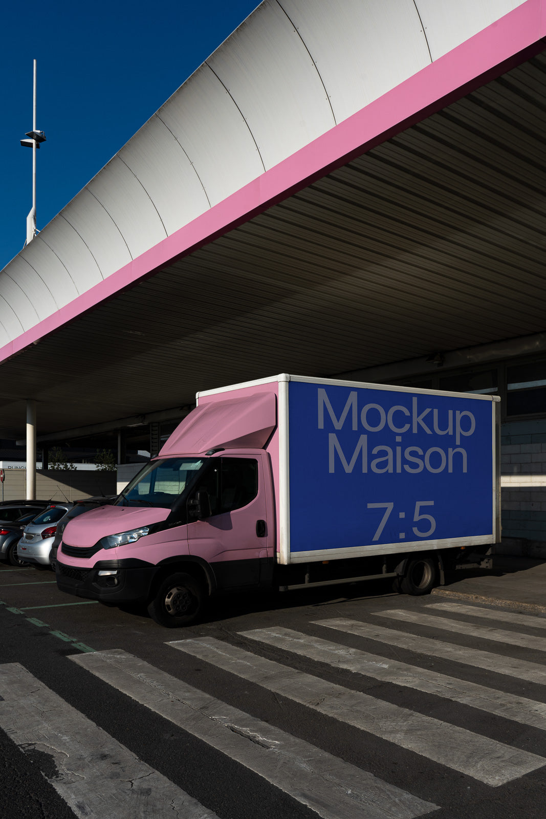 PSD mockup of a delivery truck with pink cab and blue cargo box parked under a roof, displaying the text "Mockup Maison 7:5" on the side.