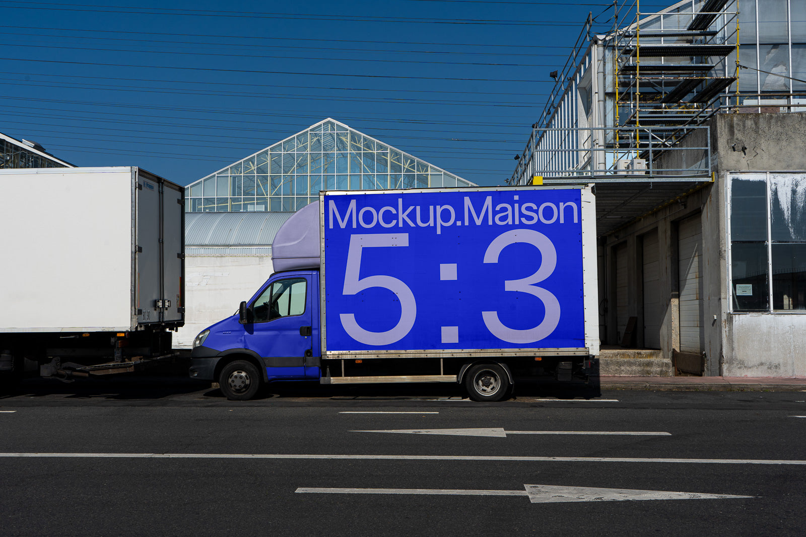 PSD mockup of a delivery truck with a large blue banner displaying the text "Mockup.Maison 5:3" parked in front of industrial buildings.