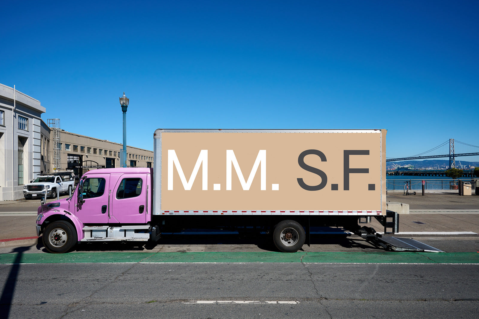 PSD mockup of a pink truck parked on a street with "M.M. S.F." in large letters on the beige cargo area, city backdrop and clear sky.
