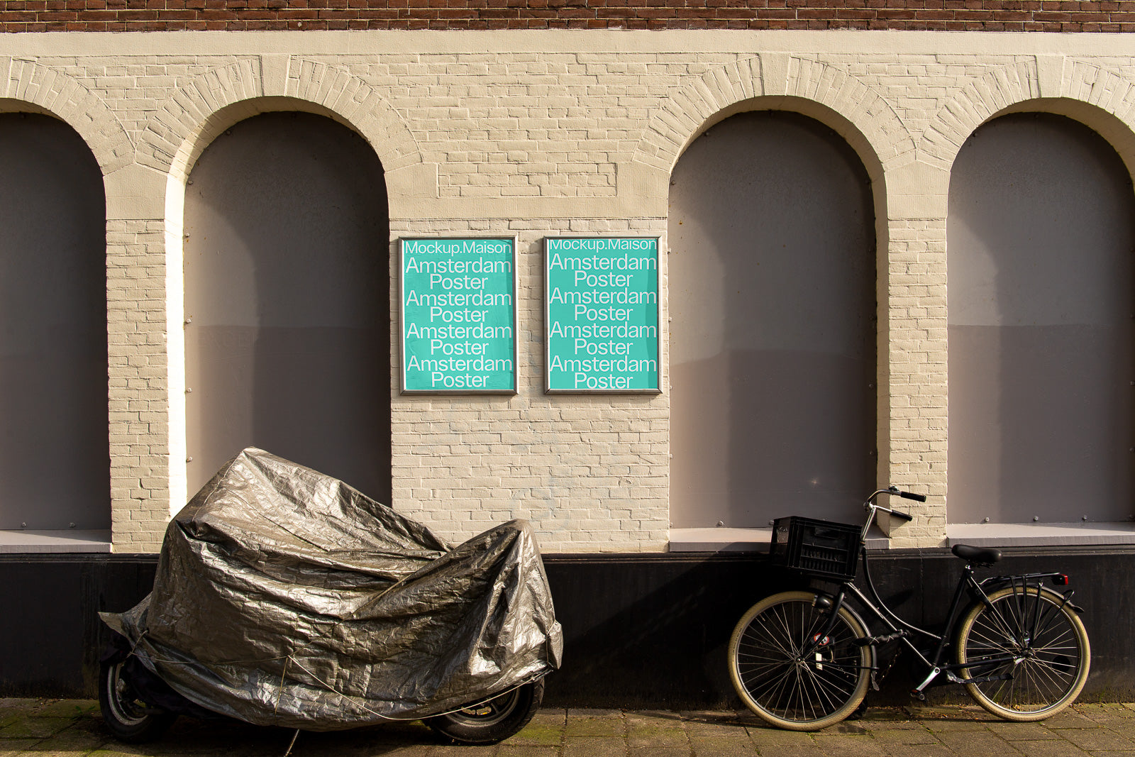 PSD mockup of outdoor poster displays against a brick wall, showing two identical teal posters with text, beside a covered motorcycle and a parked bicycle.